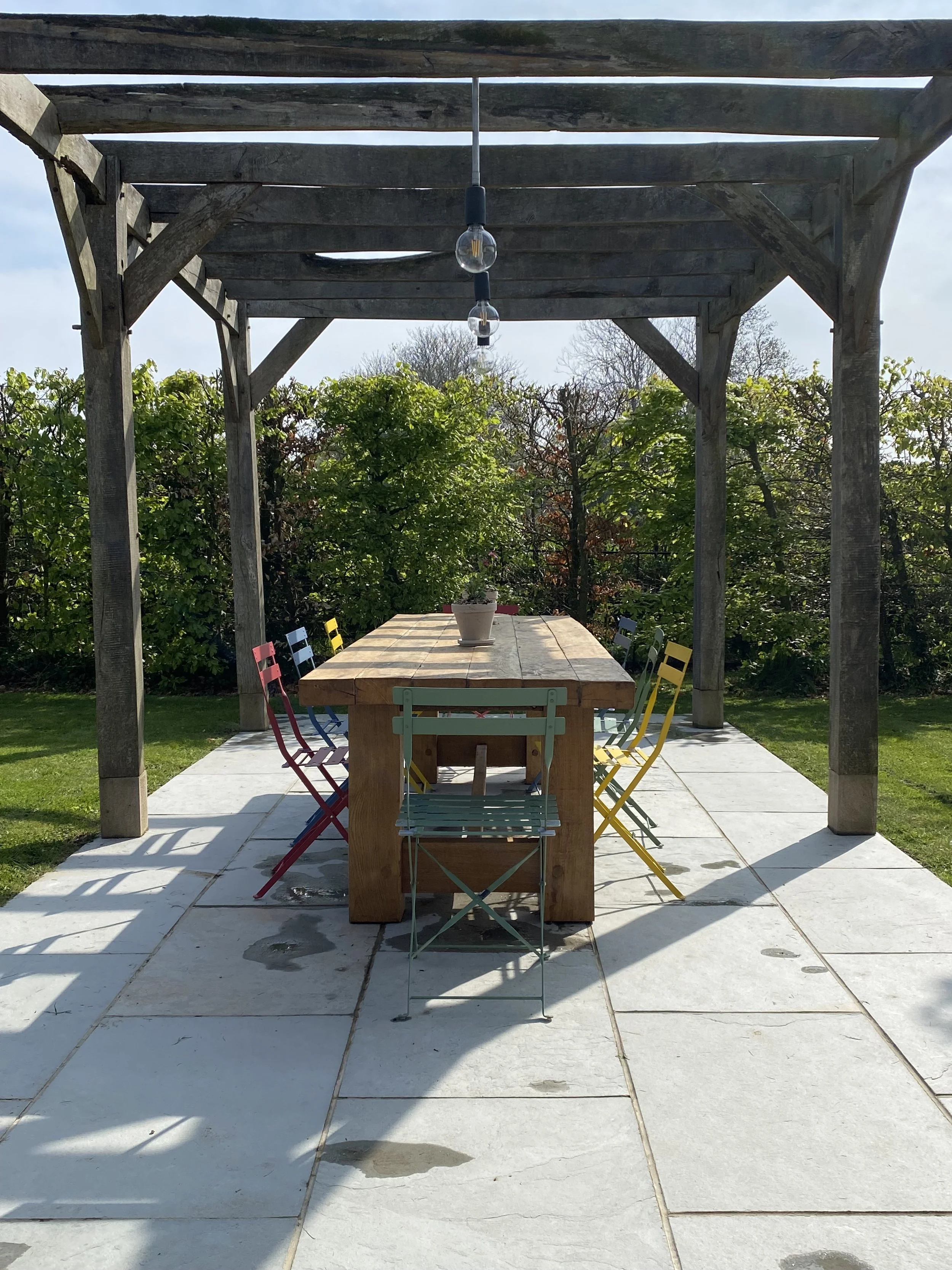 Outdoor patio with a rectangular wooden table under a wooden pergola, surrounded by colorful metal chairs, with a potted plant on the table, green bushes in the background, and a partly cloudy sky.