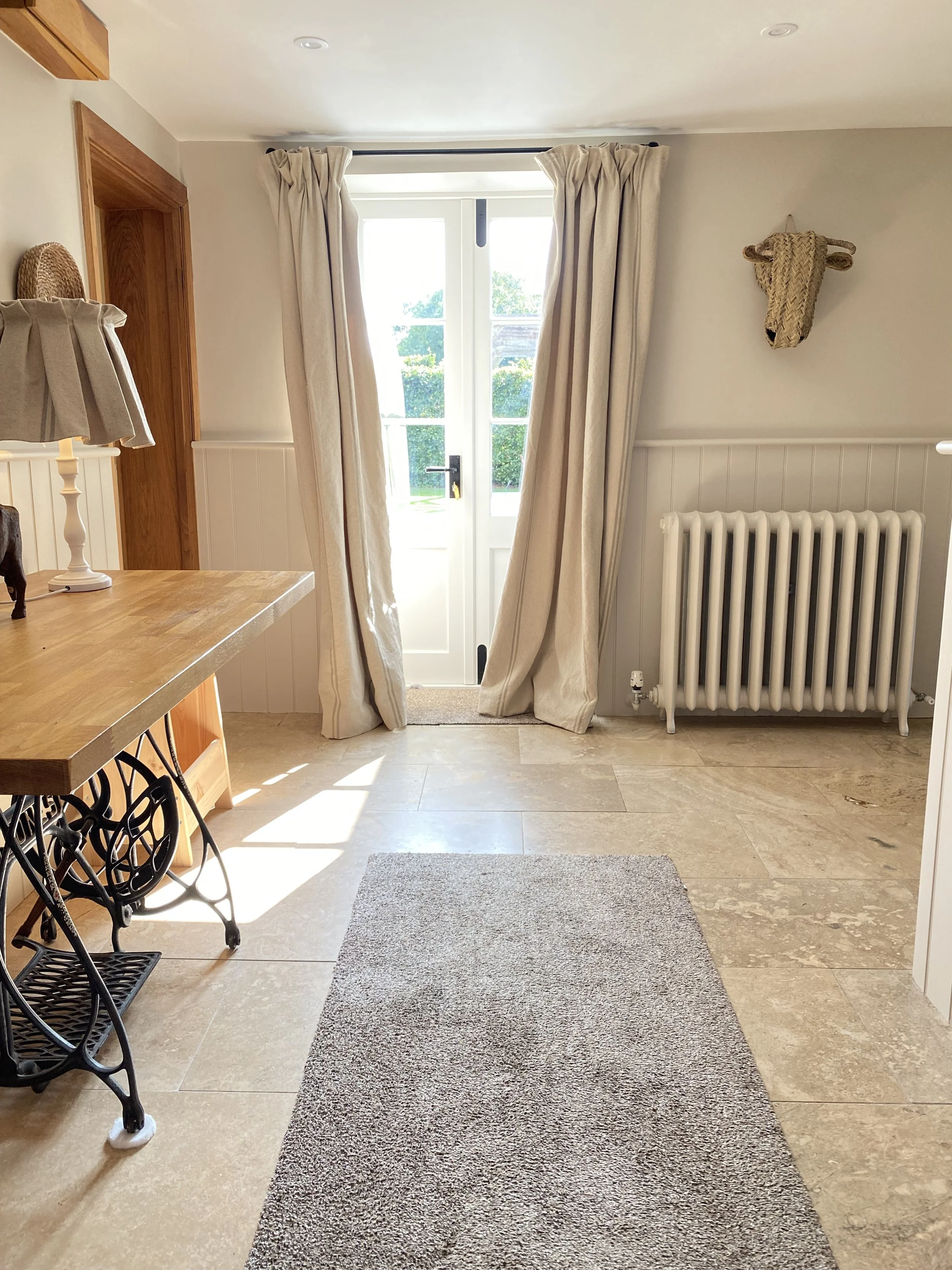 A bright dining area with beige curtains covering a glass door, a white radiator on the right, a beige and brown woven wall hanging resembling a bull's head, a wooden table on the left, and a gray rug on the tiled floor.