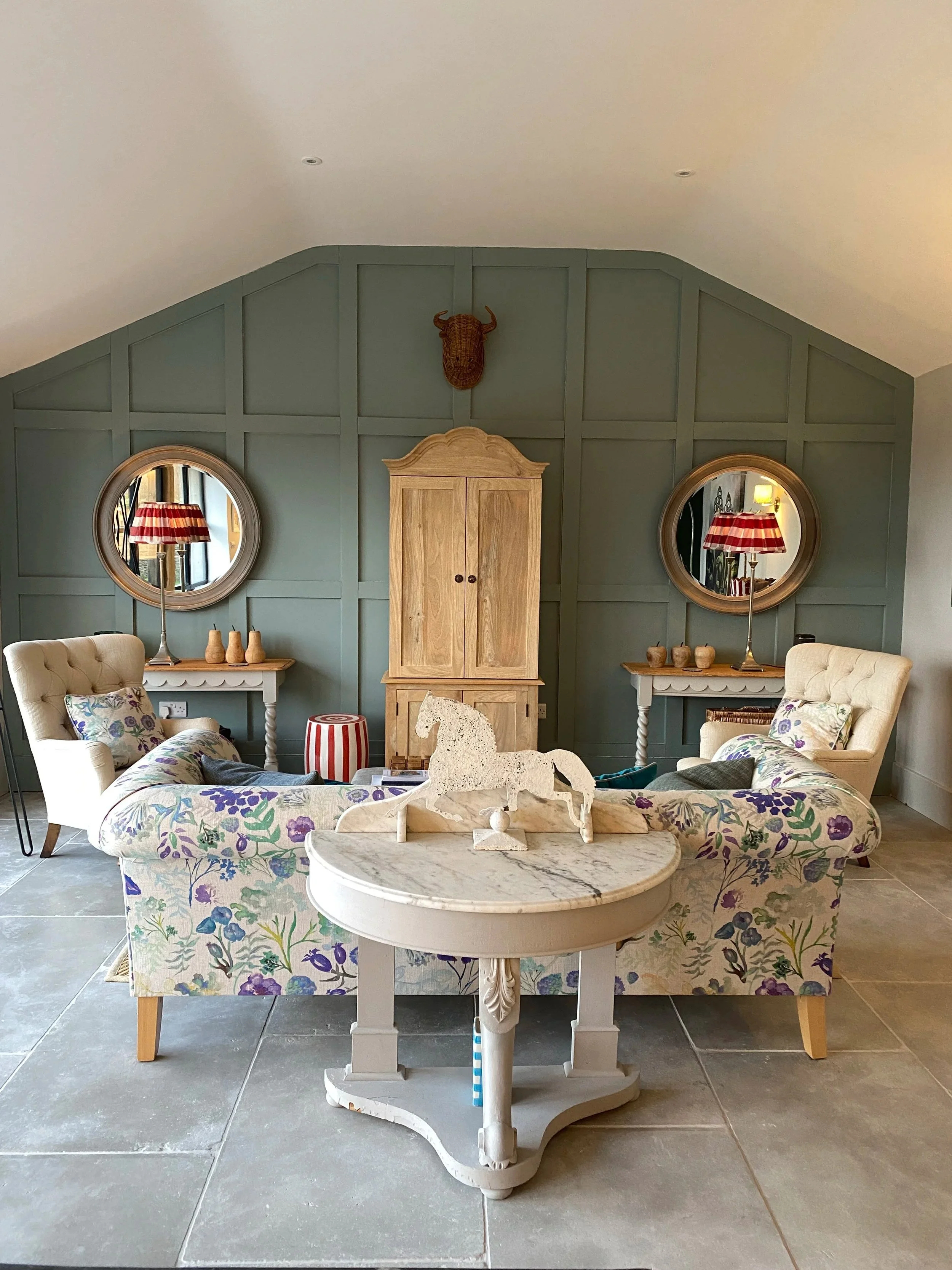 Living room with two floral-patterned sofas, a marble-top coffee table with a horse and rider sculpture, wooden side tables with lamps, an armoire, round mirrors, a table lamp with a striped shade, and a wall-mounted animal head sculpture.