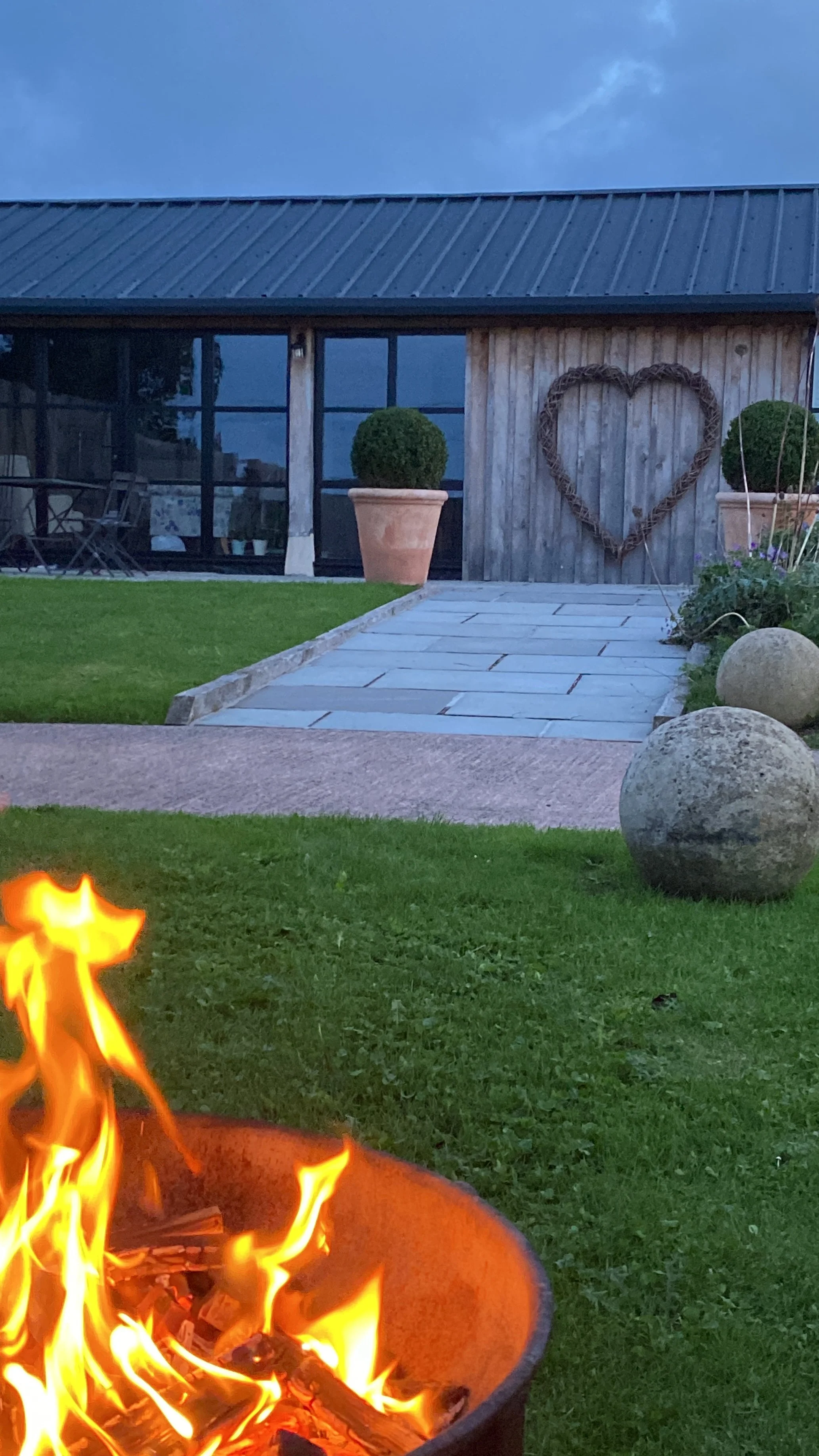 A backyard scene during dusk featuring a fire pit in the foreground, a stone pathway leading to a wooden shed with two large potted bushes and a heart-shaped wreath on the door, and a well-maintained lawn with decorative stones.
