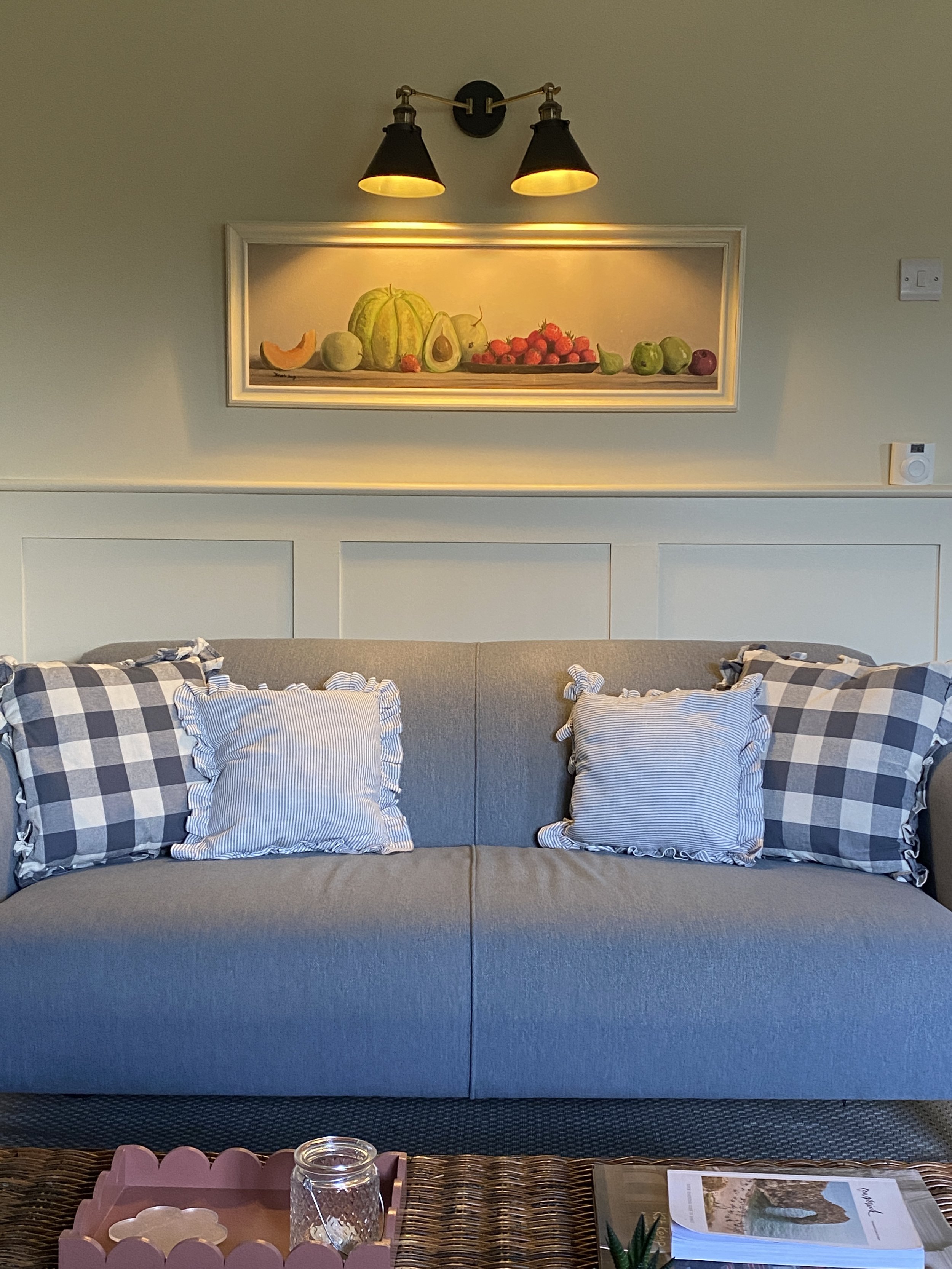 Living room with a beige sofa, checkered and striped pillows, a wicker coffee table, and a framed still life painting of fruit above the sofa, illuminated by wall-mounted lights.