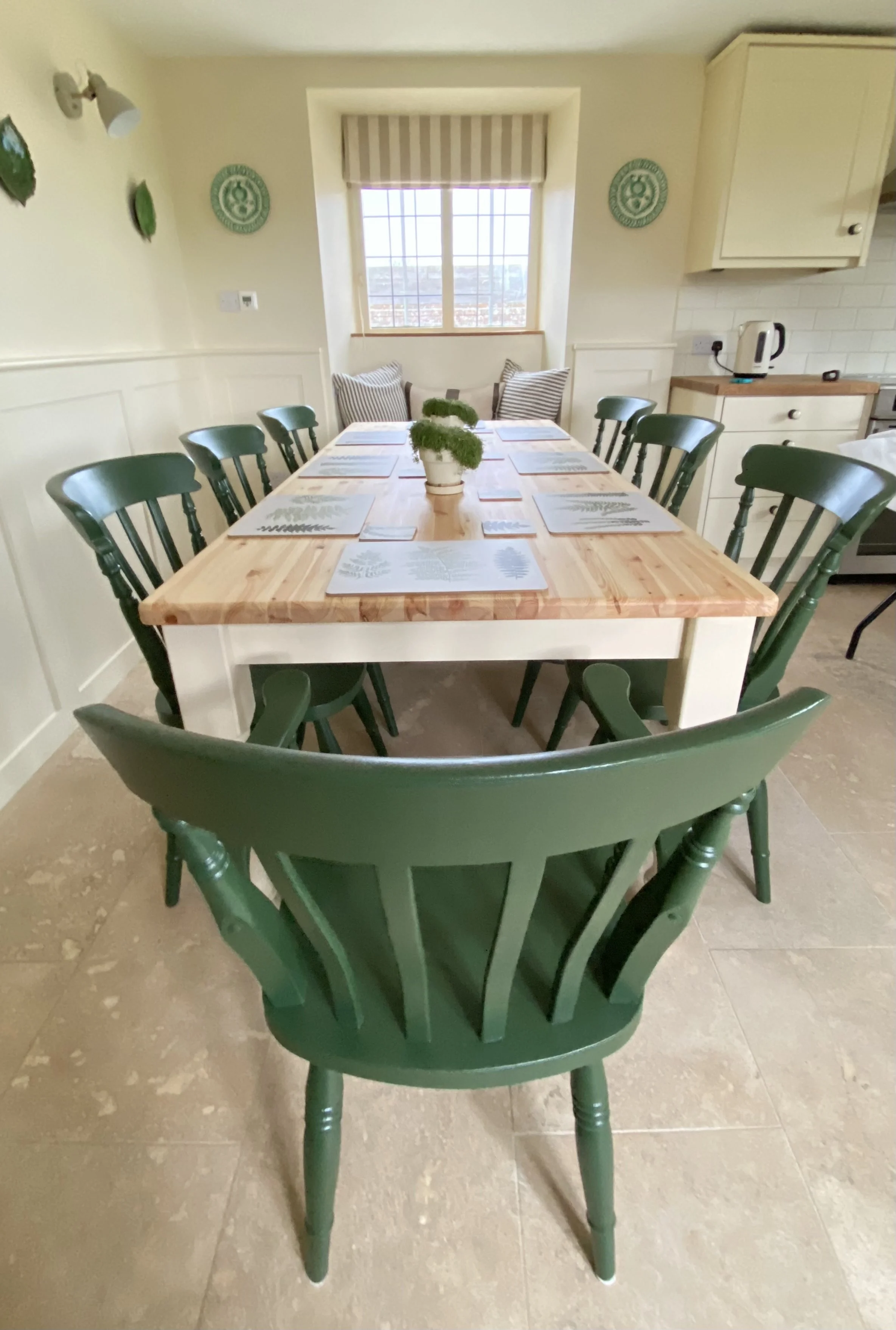 A dining room with a wooden table surrounded by twelve green chairs, set with placemats and a centerpiece of small potted plants. There is a window with a striped blind, decorative plates on the wall, and a cozy corner with cushions. Part of the kitc