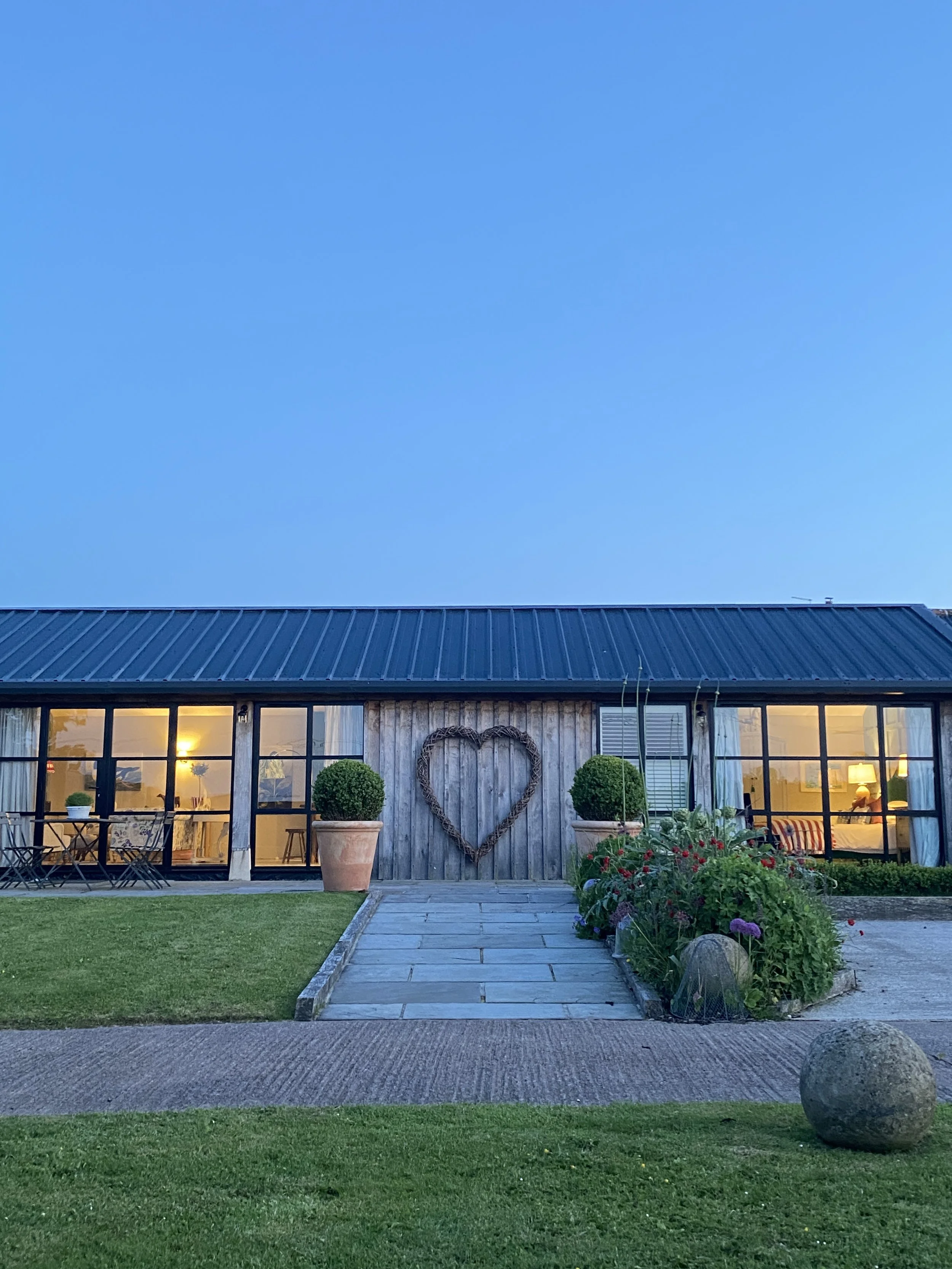 A house with large glass windows, two potted topiary plants, a heart-shaped decoration on the wooden wall, and a garden with flowers in the foreground during early evening.