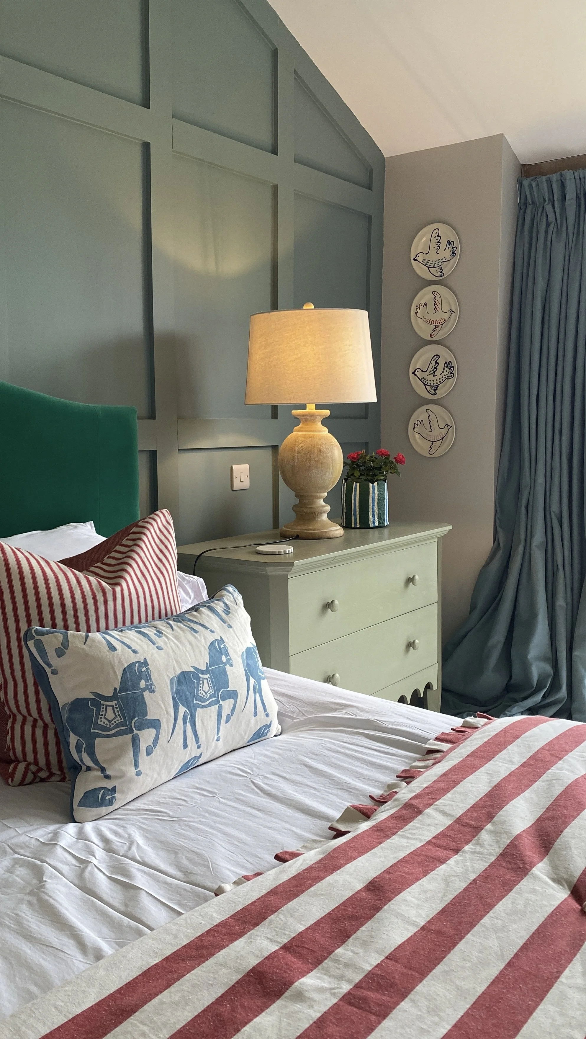 A cozy bedroom with a bed decorated with striped and solid-colored pillows, a white bedspread, and a striped blanket. A green headboard is visible, and there's a cream-colored nightstand with a beige lamp, a plant in a striped pot, and an electrical 