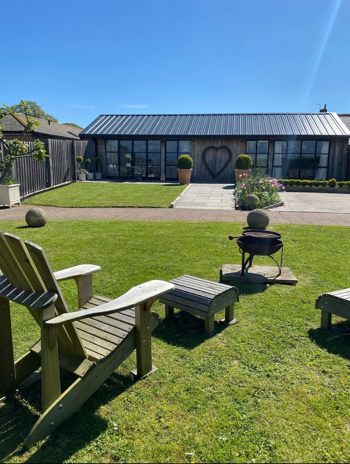 Backyard with wooden Adirondack chairs, a small fire pit, a well-maintained lawn, potted plants, and a building with large windows and a heart-shaped decoration on the wall.