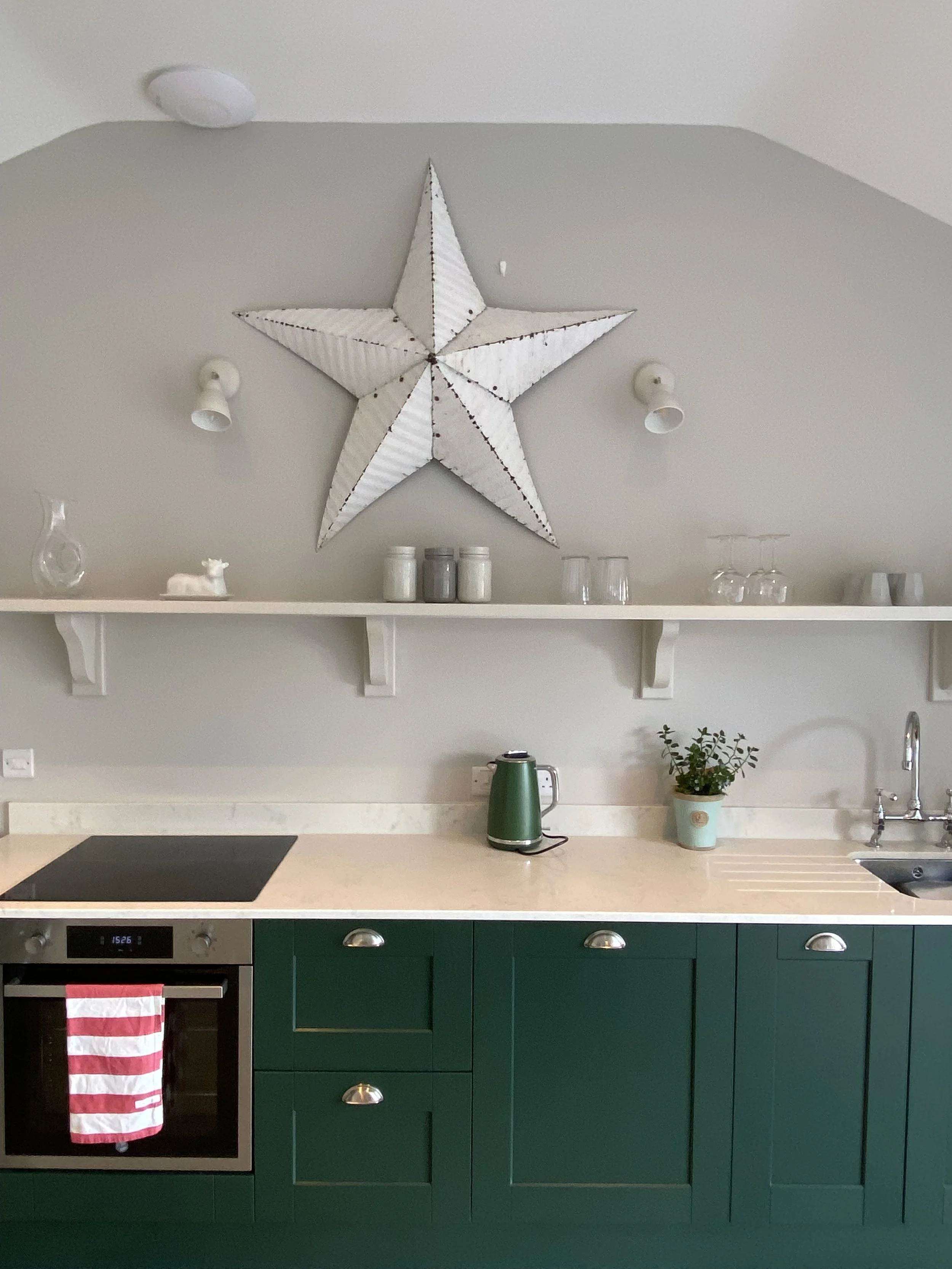 A kitchen with green lower cabinets, a beige countertop, a built-in oven with a pink and white towel hanging, a stovetop, a green electric kettle, a small potted plant, a sink, and a wall shelf above, decorated with a large white star-shaped ornament and various glassware and jars.