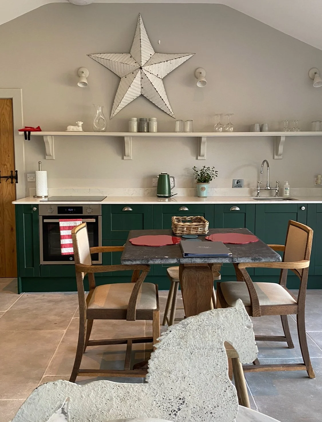 A kitchen with green cabinets, a white countertop, and a wooden table with chairs. Decor includes a large star on the wall and a horse-shaped decor piece in the foreground.