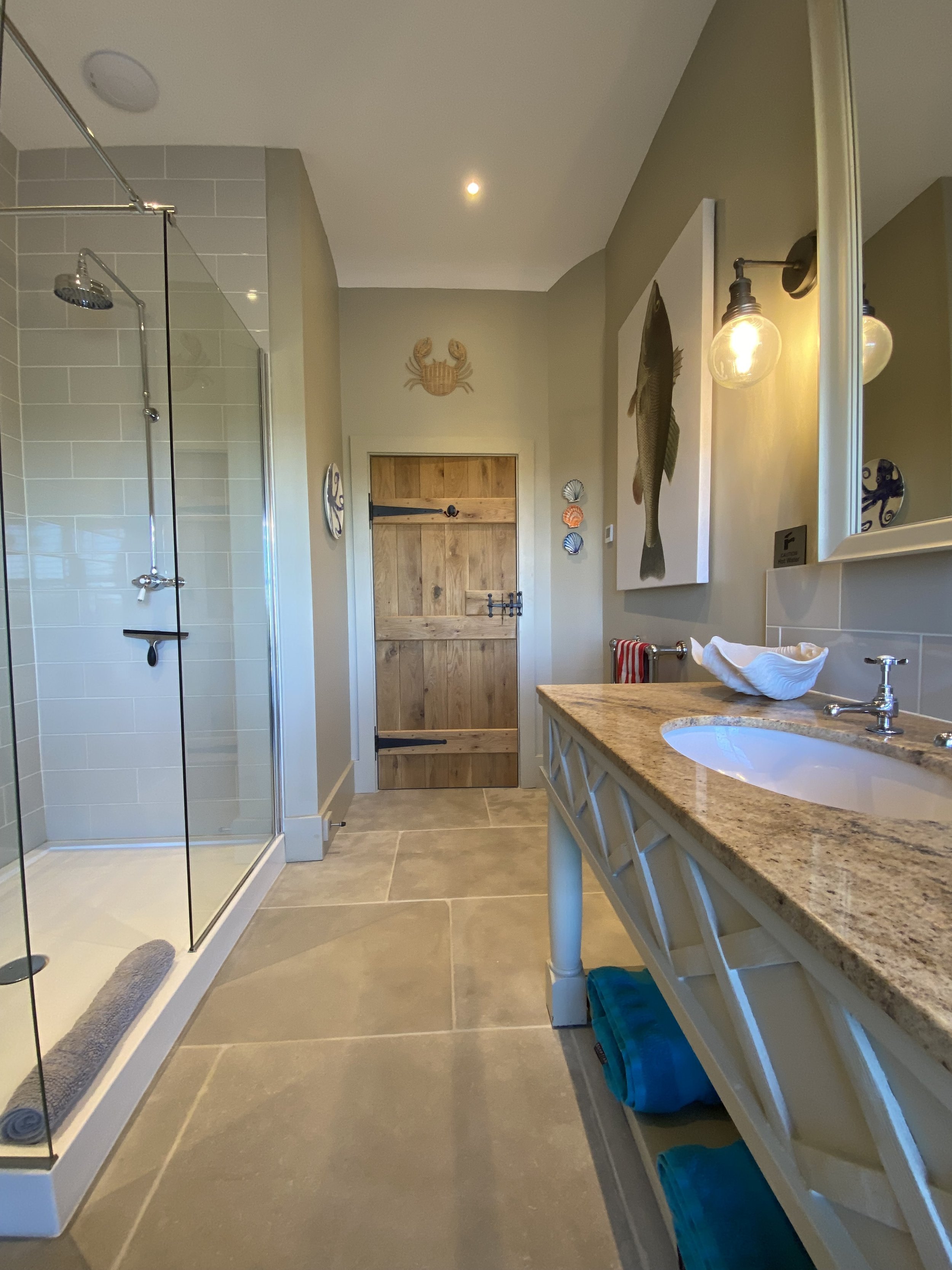 A bathroom with a walk-in shower on the left, a wooden door on the back wall, and a granite countertop sink on the right with a large mirror, mounted lights, and decorative sea shell artwork.