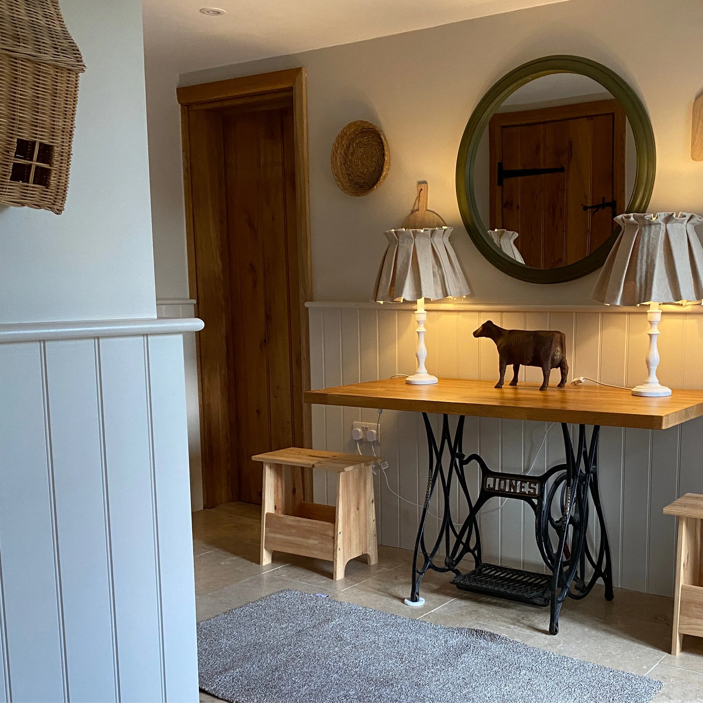 Interior view of a cozy room featuring a wooden door, a large round mirror, two table lamps with pleated shades, a small cow figurine on a wooden table, wicker basket wall decor, and light-colored paneling on the lower half of the wall.
