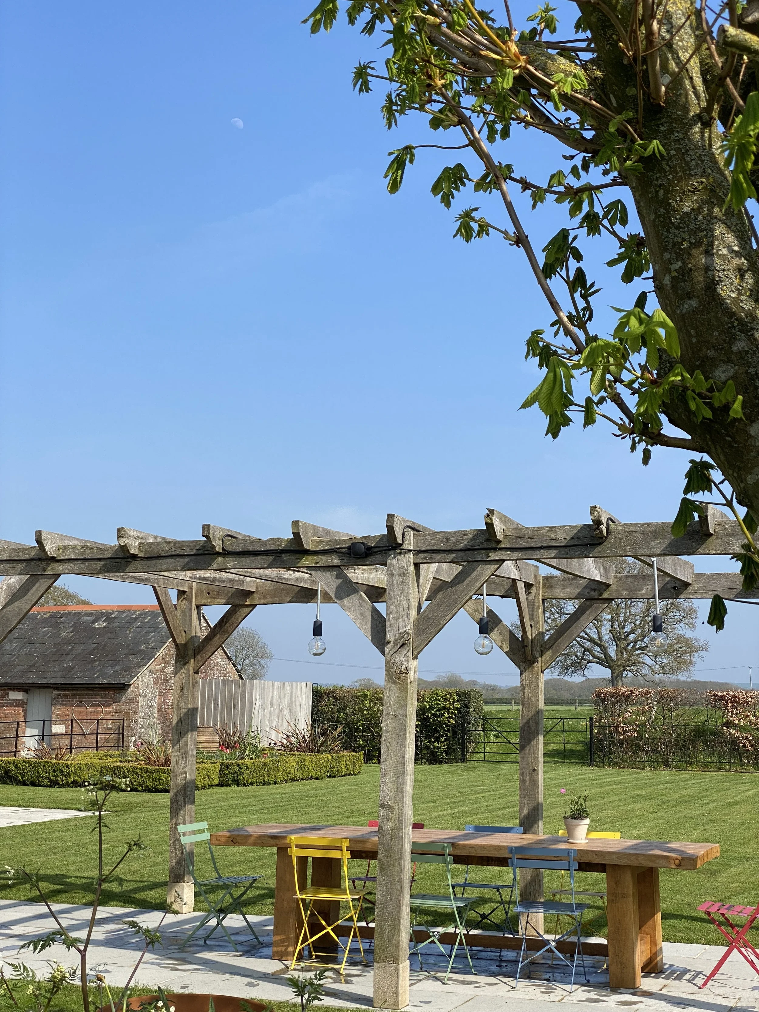 An outdoor garden patio with a wooden pergola, hanging light bulbs, a picnic table, colorful chairs, a potted plant, and a well-manicured lawn with a building and trees in the background, under a clear blue sky with a visible moon.