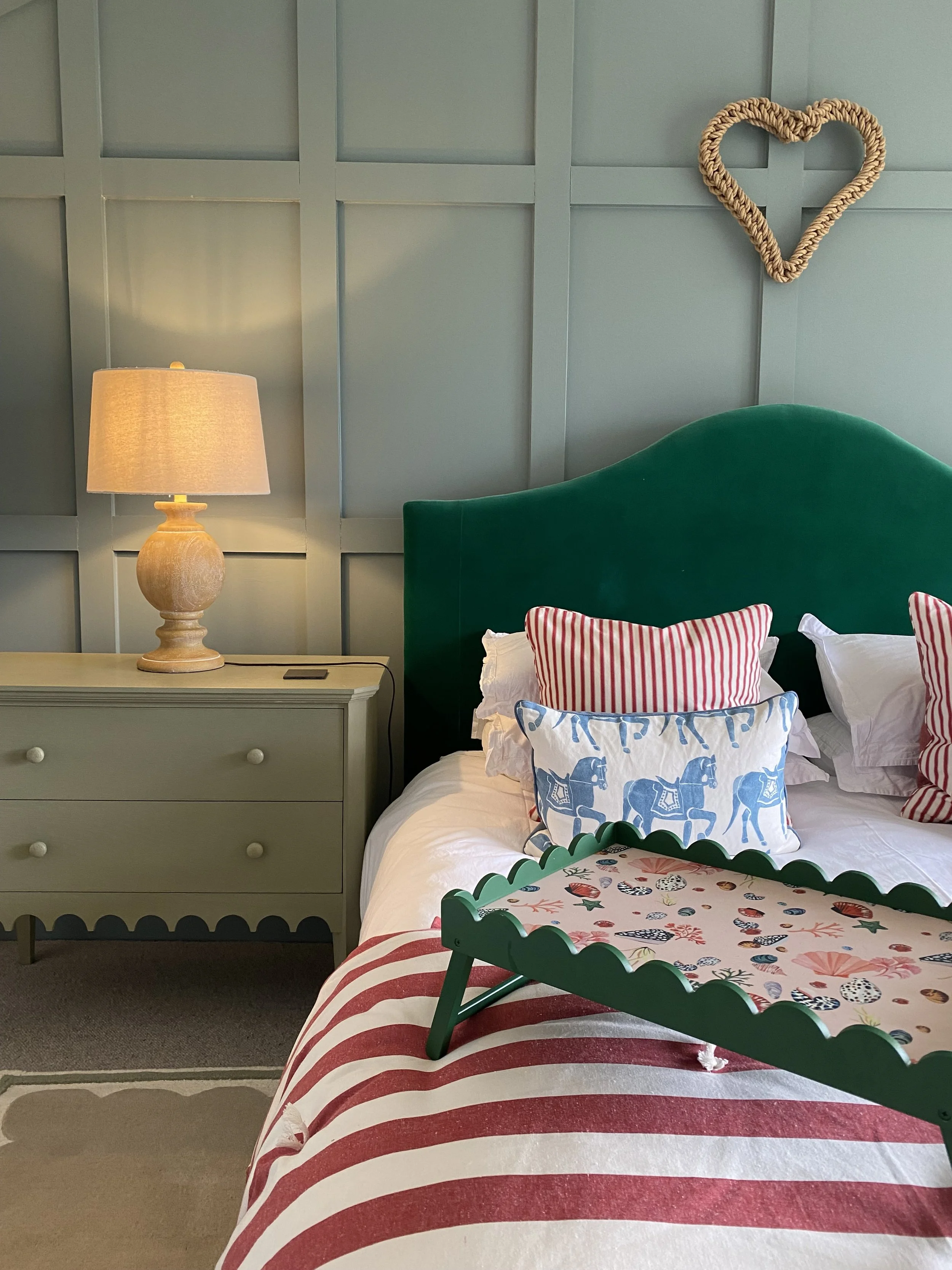 Bedroom with green headboard, striped pillows, decorative tray, nightstand with lamp, heart-shaped wall decoration.