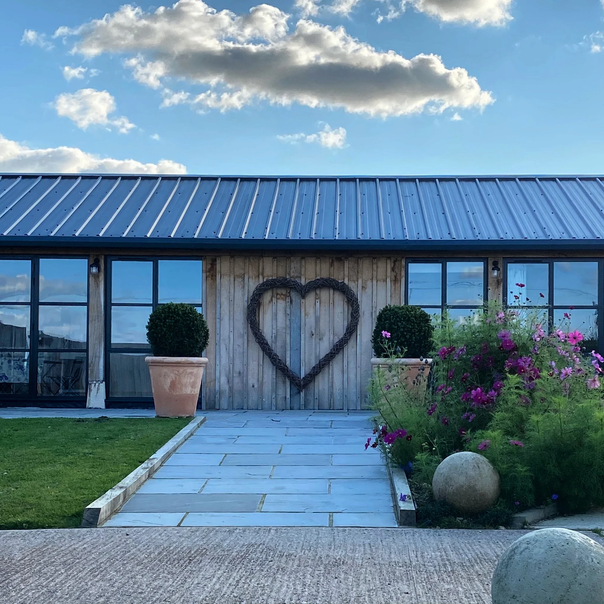 A garden pathway leading to a wooden building with large windows and a metal roof, decorated with a heart-shaped wreath on the wall and pink and purple flowers on the right side.