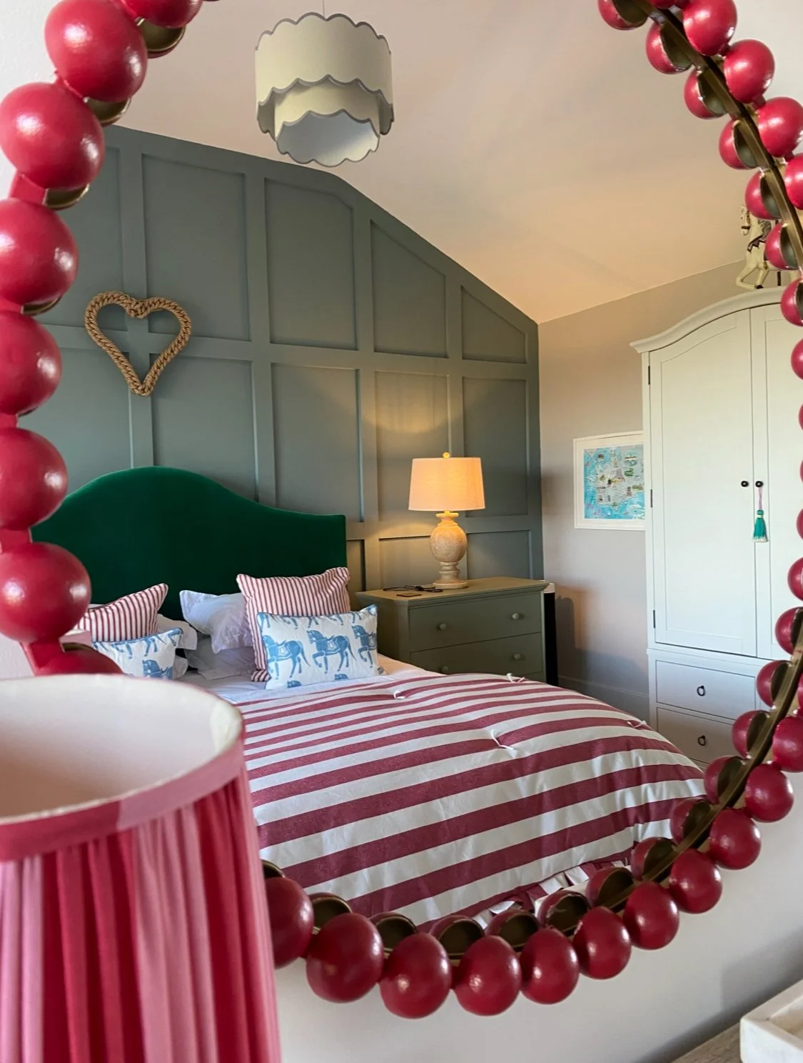 Bedroom with a bed that has a green headboard, striped red and white bedding, and pillows with horse and striped patterns. There is a nightstand with a lamp and a framed map on the wall. The photo is taken through a mirror decorated with red beads and a pink lampshade.