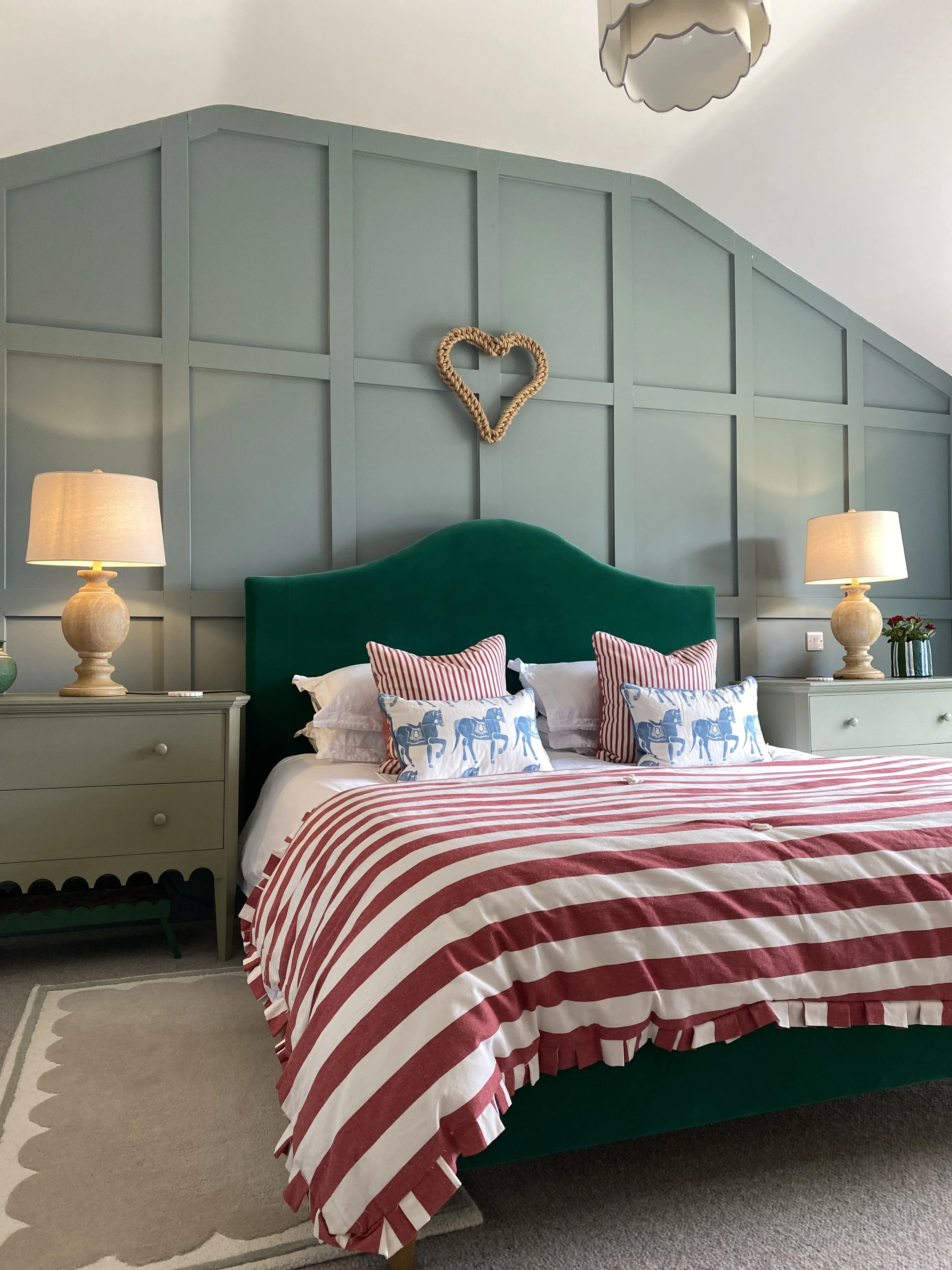 A cozy bedroom features a green velvet headboard, striped red and white bedding, and decorative pillows with horse and striped patterns. Two beige bedside tables with lamps and decorative items are on either side of the bed. The wall behind the bed i