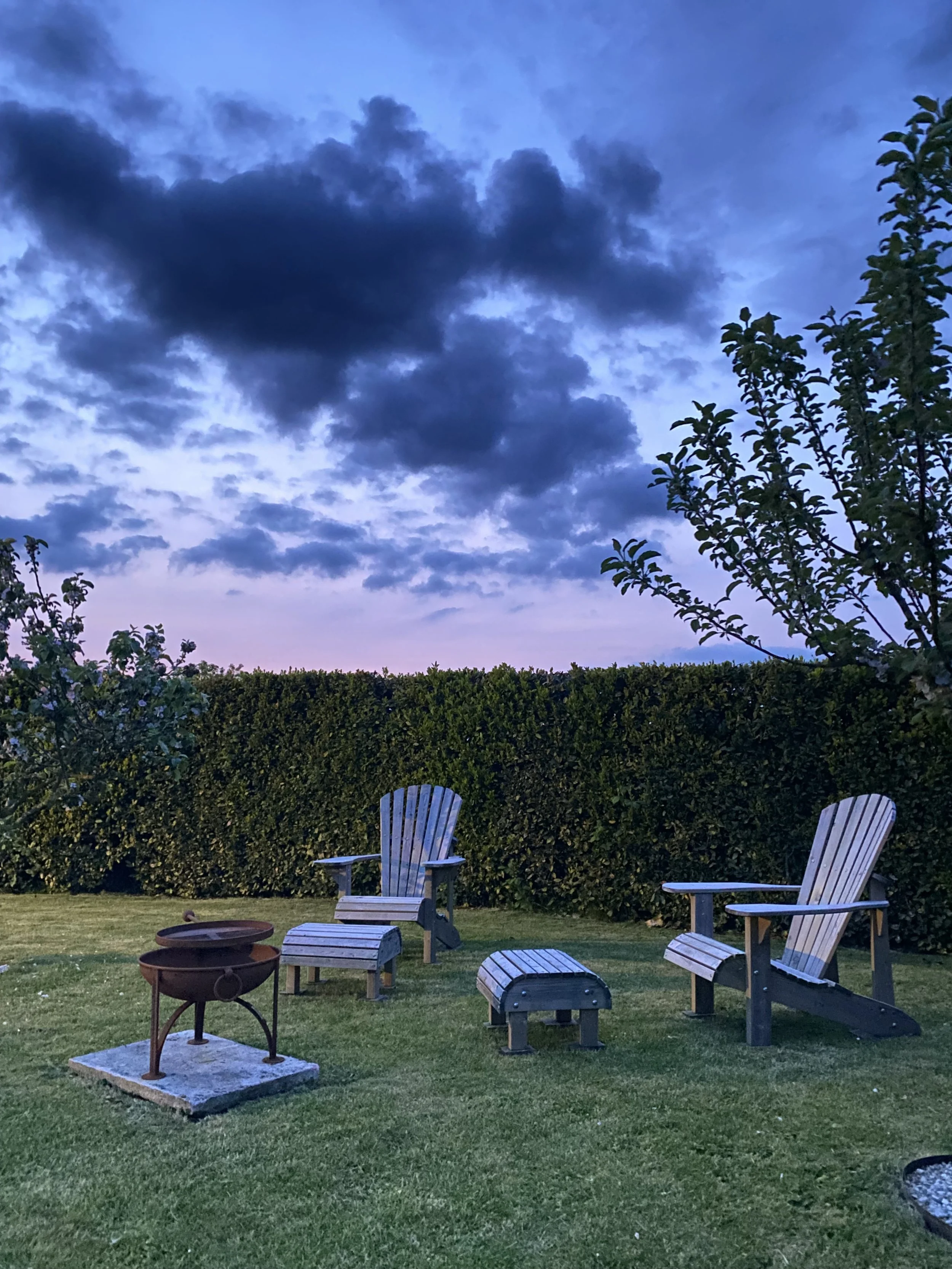Evening backyard scene with two Adirondack chairs, a fire pit, and a hedge against a purple and blue sky with dark clouds.