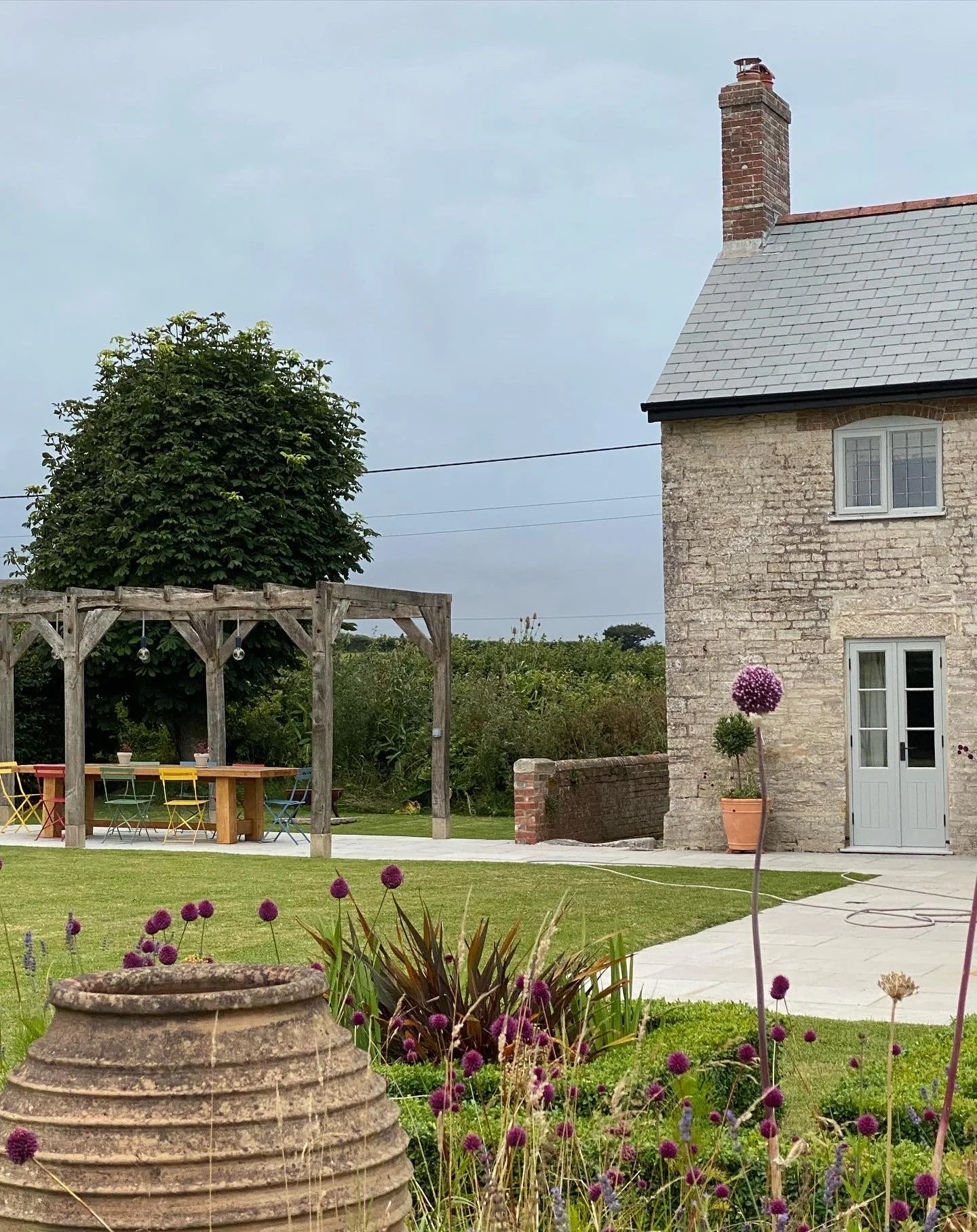 A stone house with a chimney and white-framed windows, a potted plant with purple flowers, a neatly trimmed lawn, and a garden with purple flowers. There is a wooden pergola with string lights, outdoor dining table, and chairs on a patio, and a cloud
