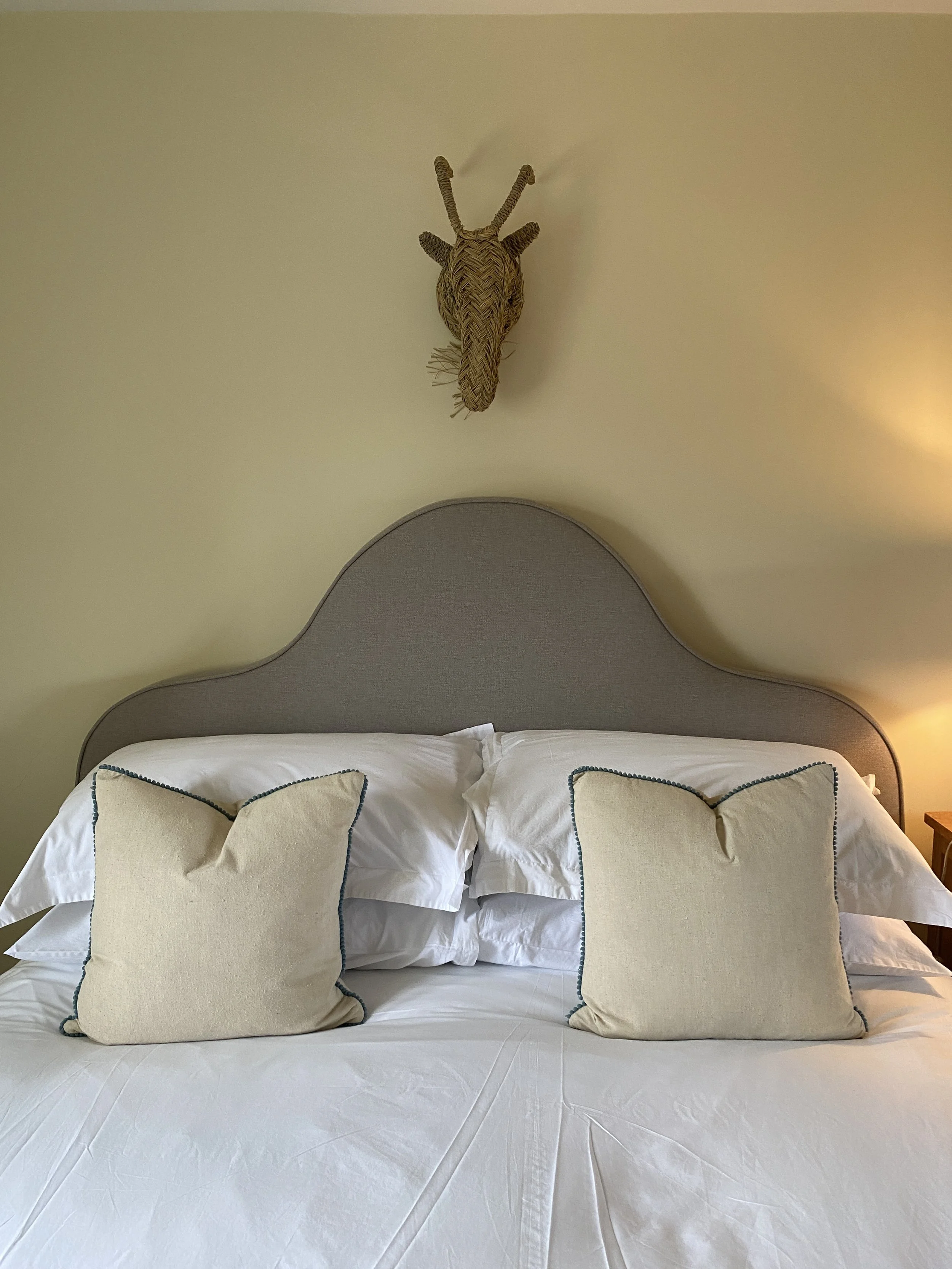 A neatly made bed with two white pillows and two beige throw pillows with blue piping. There is a beige, curved headboard and a yellow wall behind the bed. A wicker animal head is mounted on the wall above the headboard.