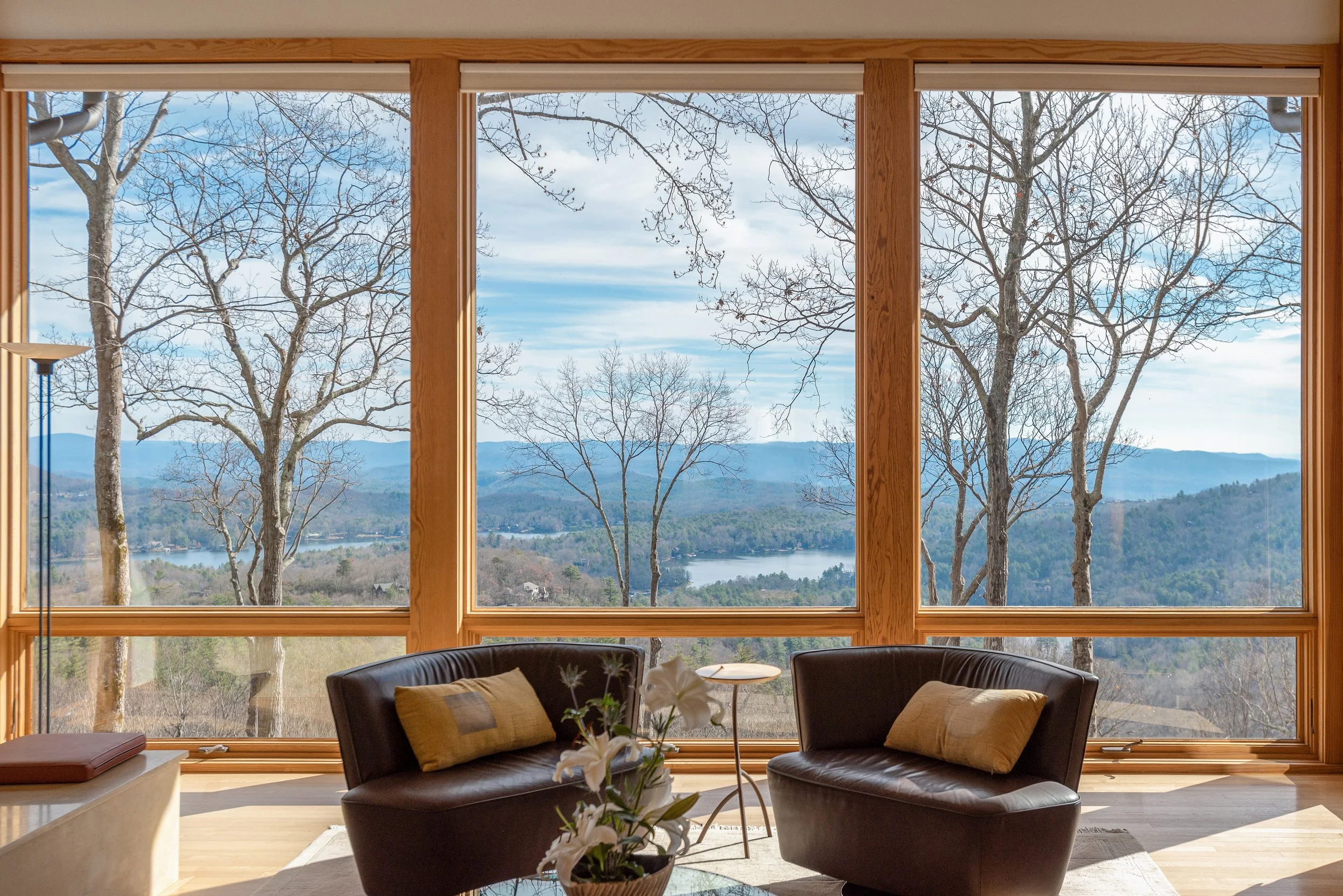 Living room with large wooden-framed window overlooking snow-covered trees and a distant lake, with chairs and a side table inside.