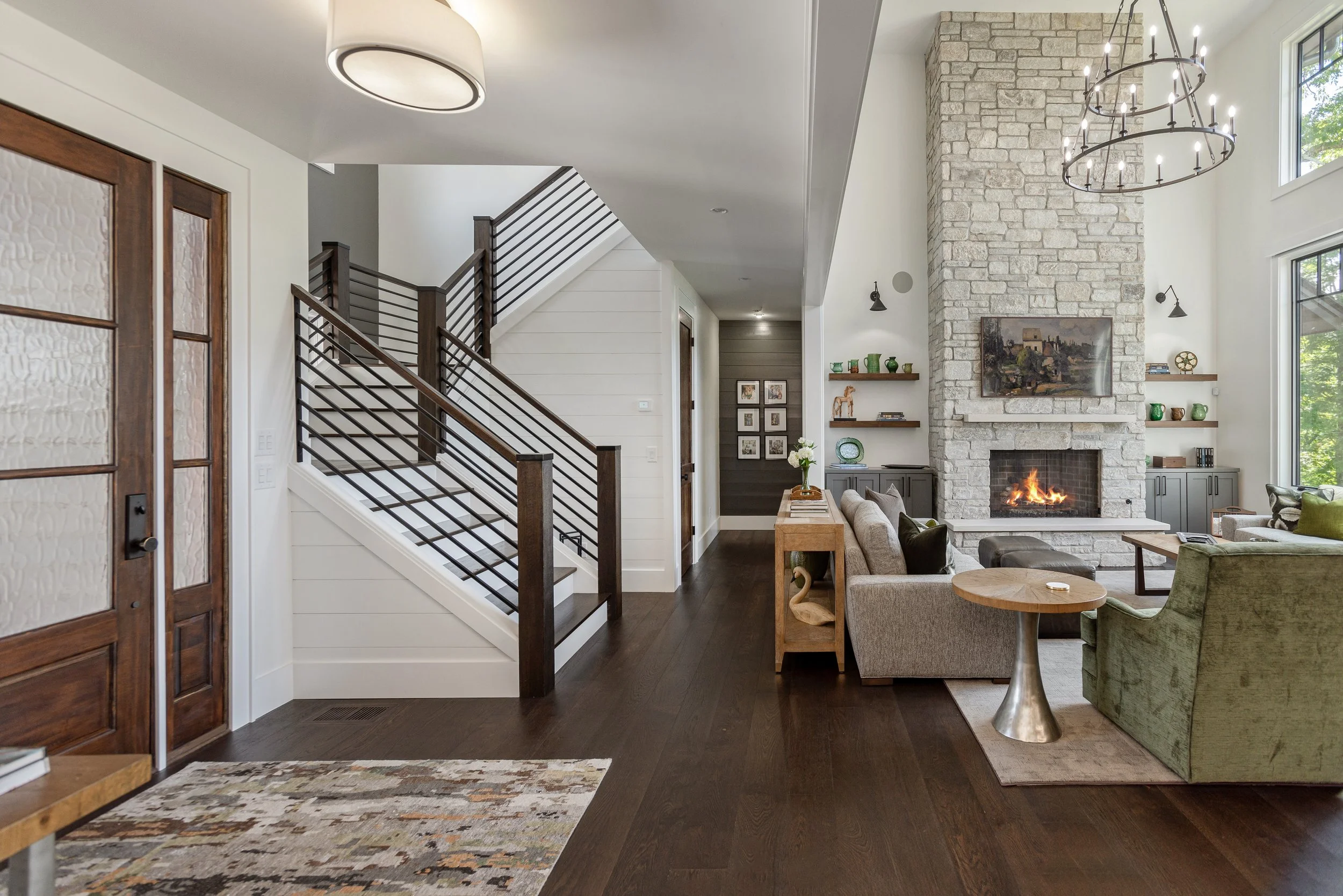 Living room with stone fireplace, modern chandelier, wall-mounted lights, and a staircase with dark wood and metal railing.
