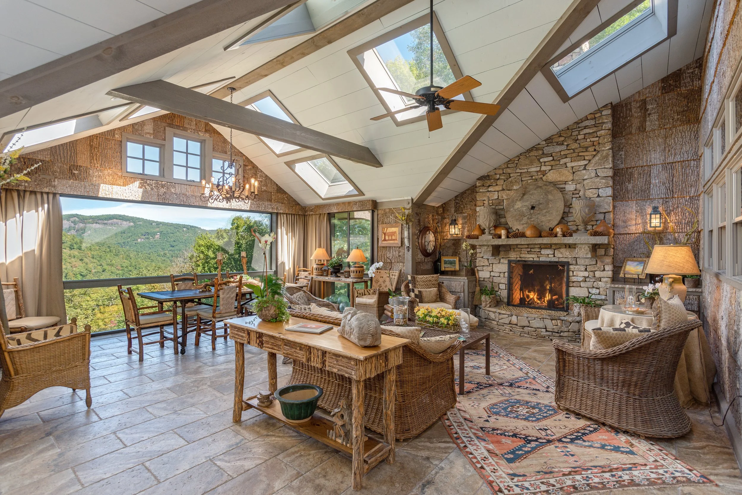 A spacious living room with large windows showcasing a mountain view, a stone fireplace with decorative pottery, rustic wooden furniture including a dining table and chairs, a ceiling fan and skylights, and a colorful area rug.