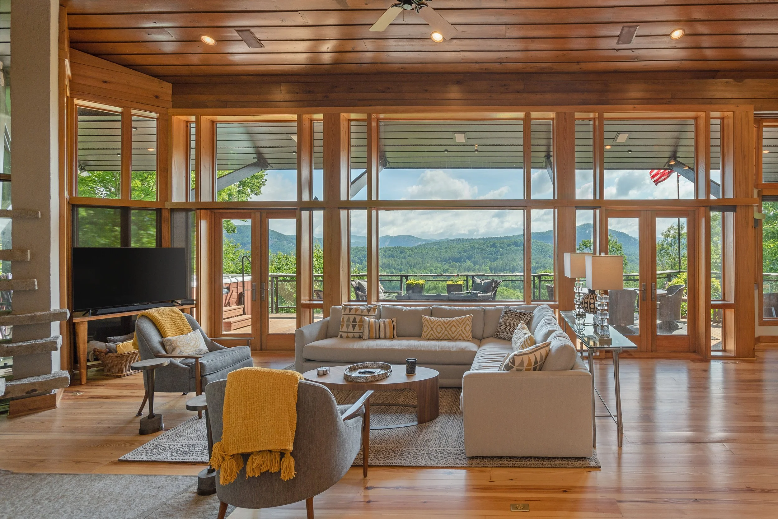 Interior of a modern living room with large windows showcasing mountains and trees outside. The room features a cream sofa with decorative pillows, a glass-top table, and a gray armchair with a yellow throw blanket. There is a flat-screen TV on a sta