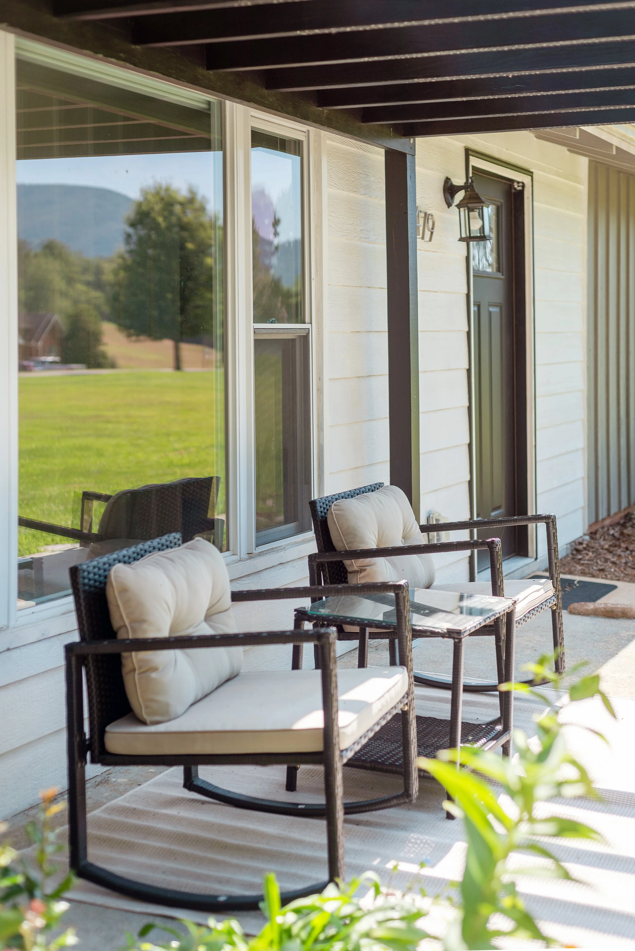 Patio with black wicker chairs and beige cushions, glass-top tables, sliding glass door, exterior wall with house number, and a porch light fixture, overlooking a green lawn and distant hills.