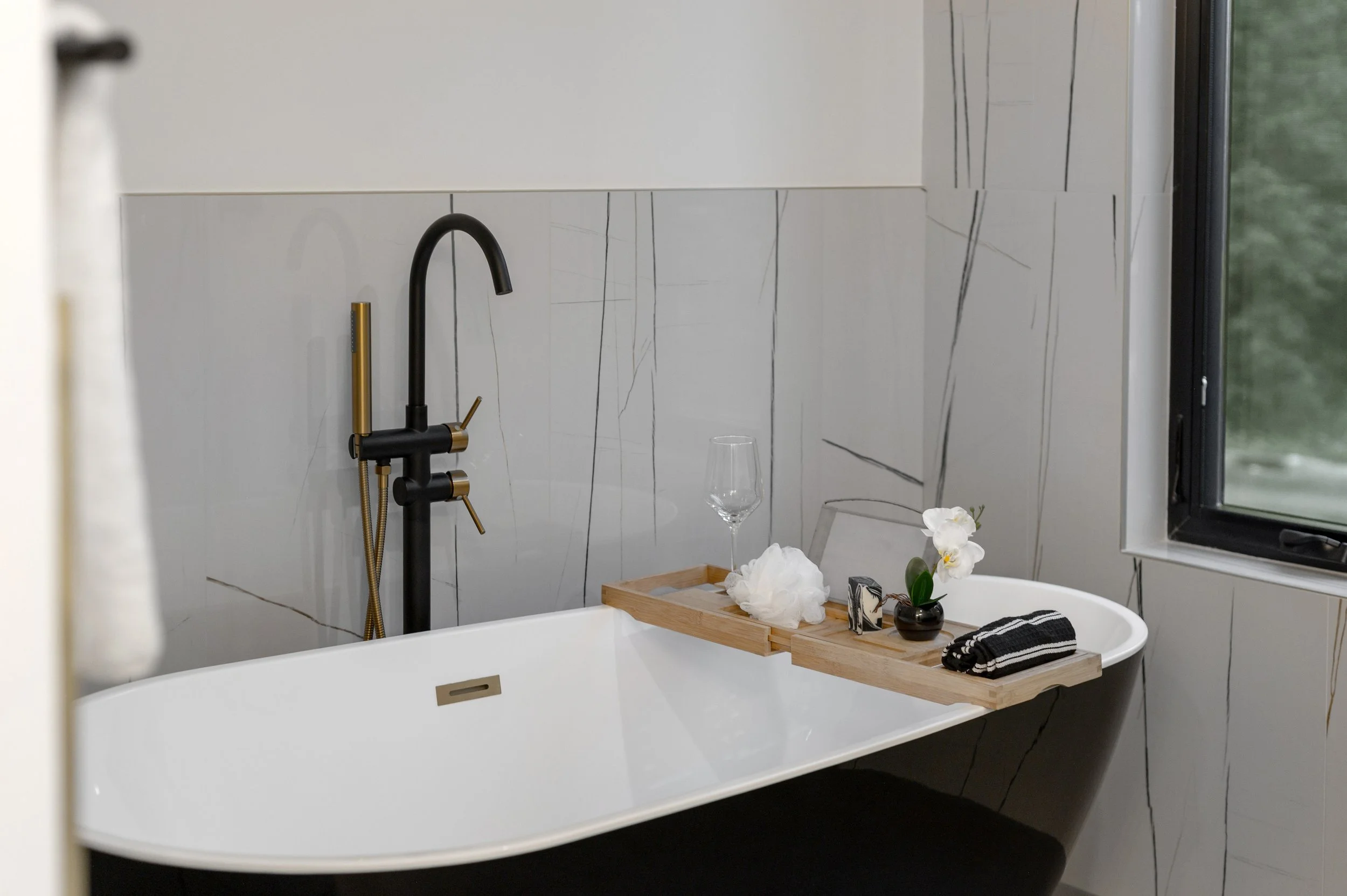 Modern bathroom with a white free-standing bathtub, black and gold faucet, and a wooden bath tray holding towels, flowers, and toiletries. A window to the right allows natural light.