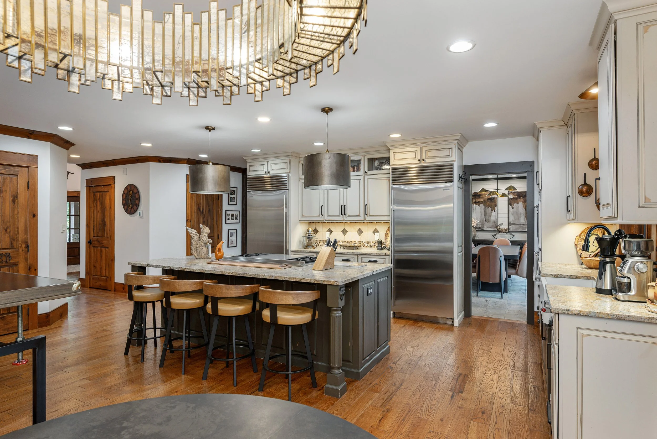 A spacious kitchen with white cabinets, stainless steel appliances, a central island with a granite countertop, and a wooden floor. Modern pendant lights hang above the island, and a large decorative chandelier is on the ceiling.