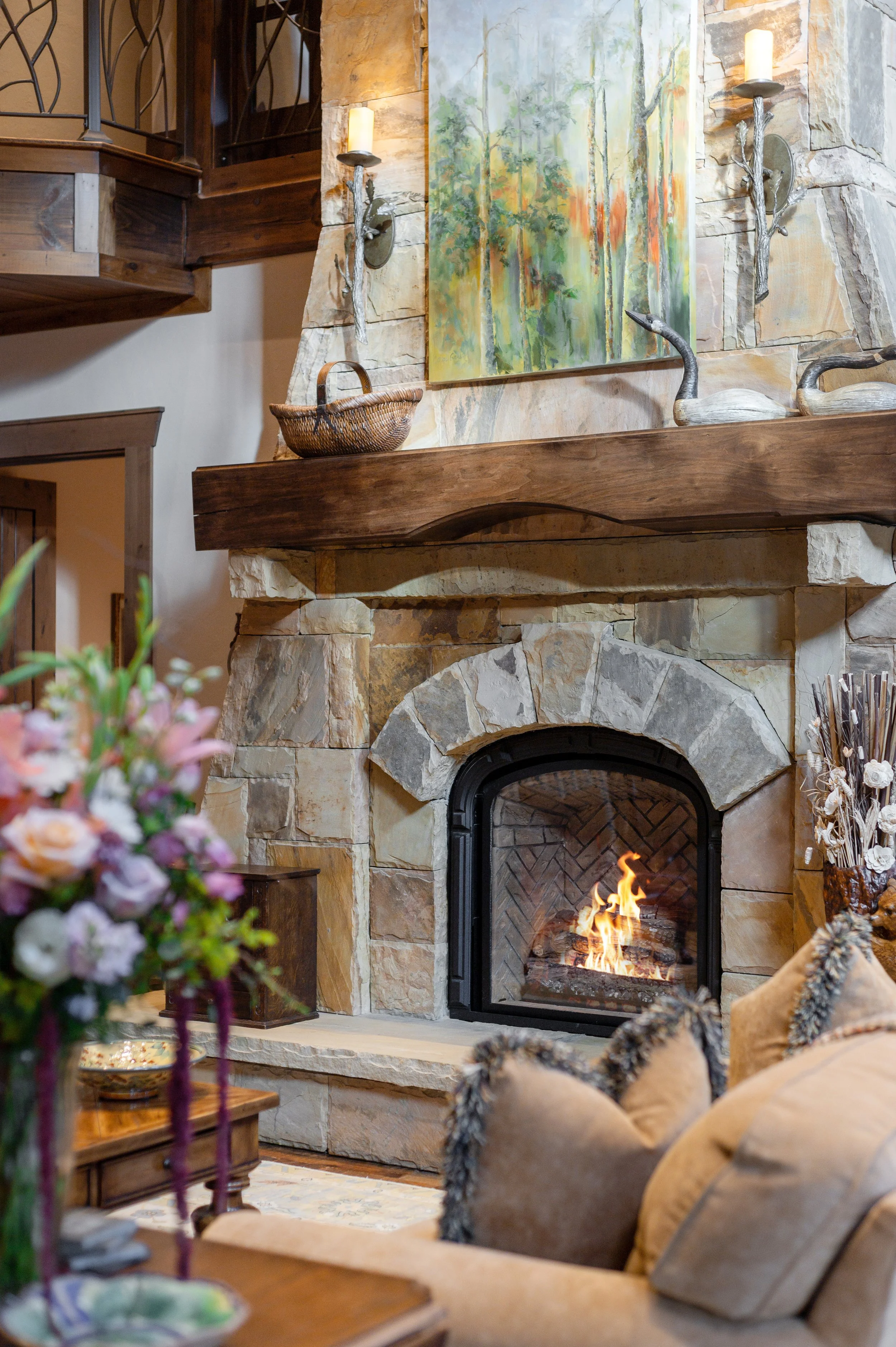 A cozy living room features a stone fireplace with a burning fire, a wooden mantel, and decorative sculpted ducks. A colorful abstract painting hangs above the fireplace. In the foreground, part of a floral arrangement and a wooden table with a bowl 