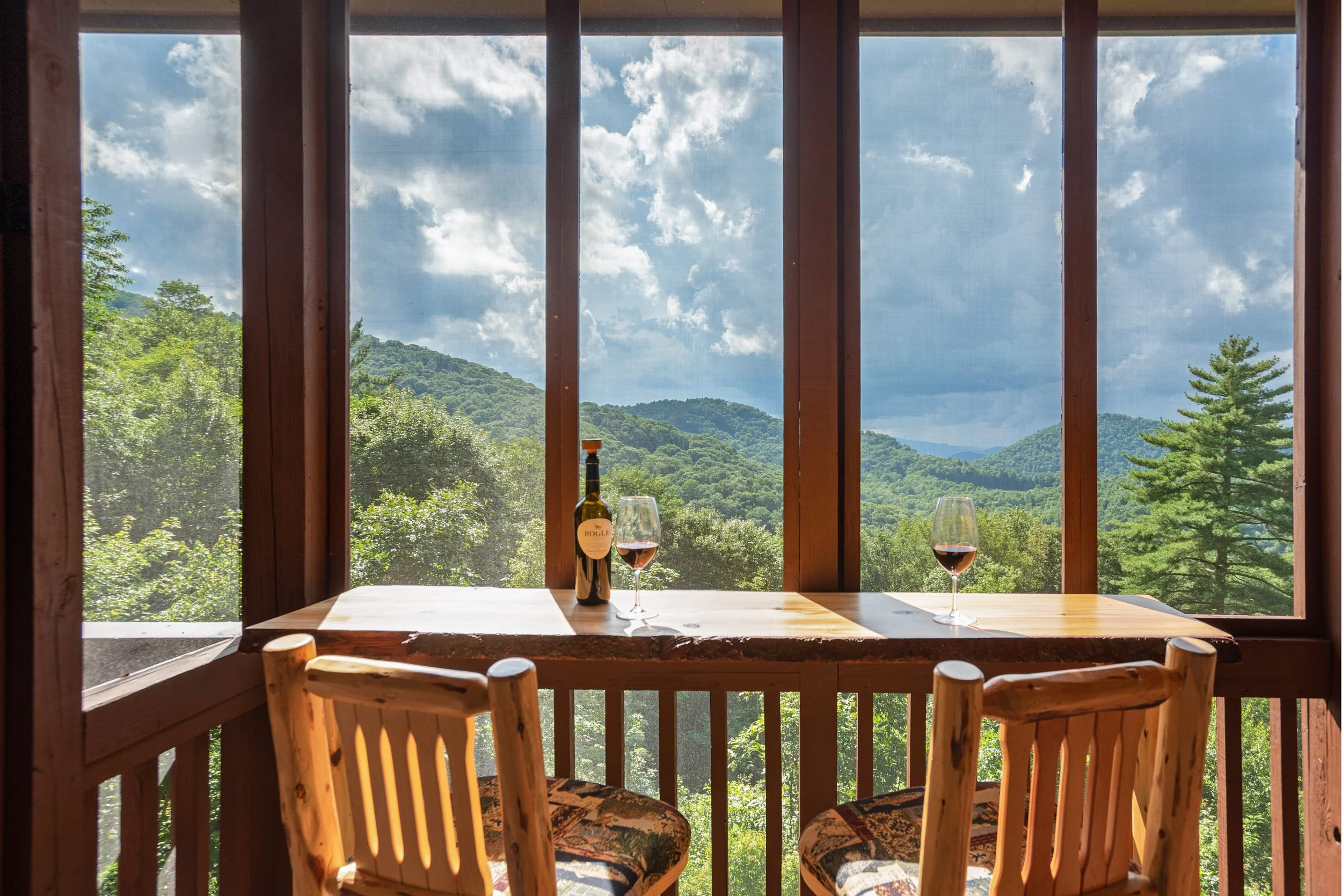 View from inside a wooden cabin with a large window showing green trees and distant mountains under a partly cloudy sky. On the table are a bottle of red wine and two glasses of red wine.