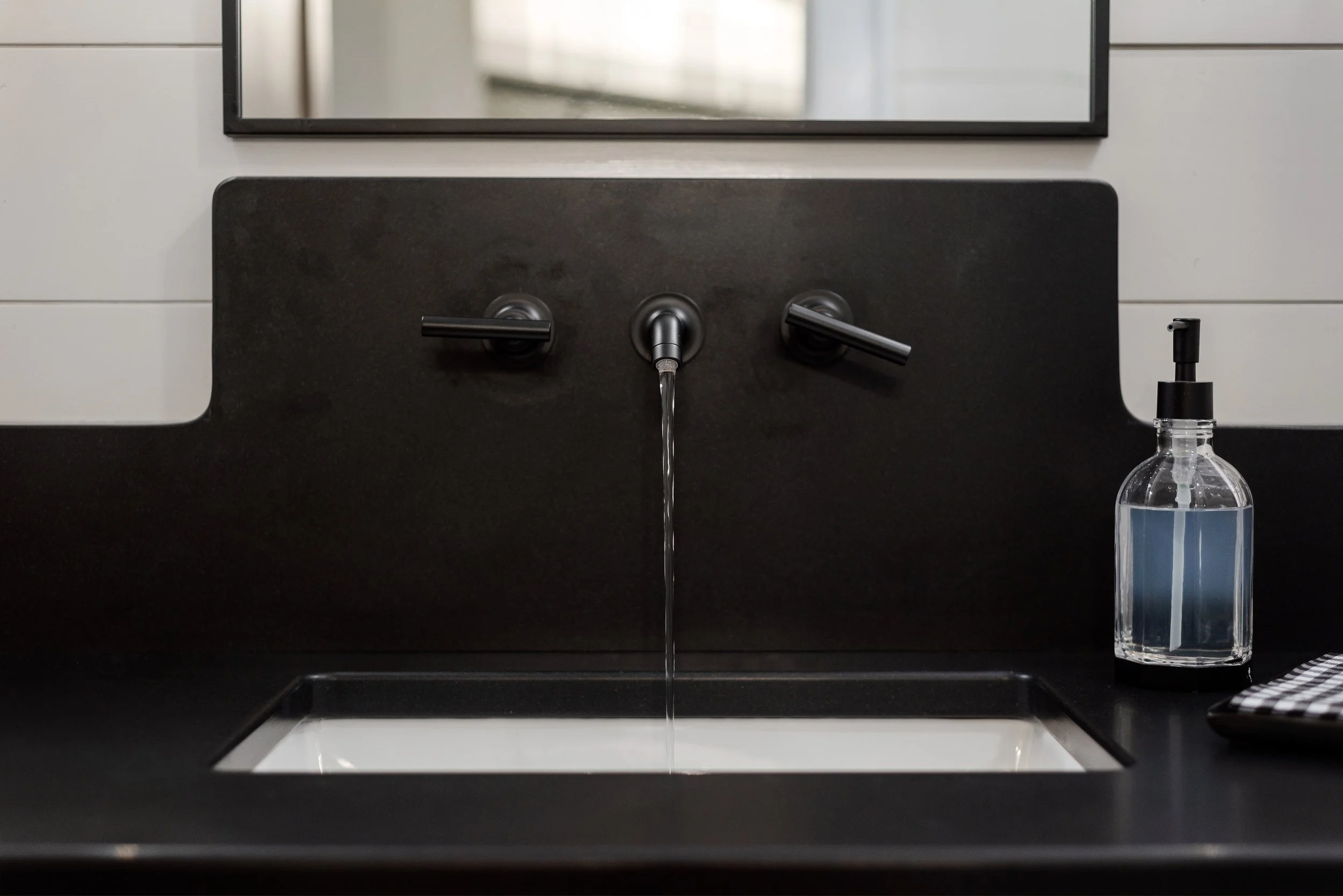 A modern kitchen sink with a black countertop, a faucet with water running, a mirror above the sink, and a bottle of hand sanitizer on the right side.