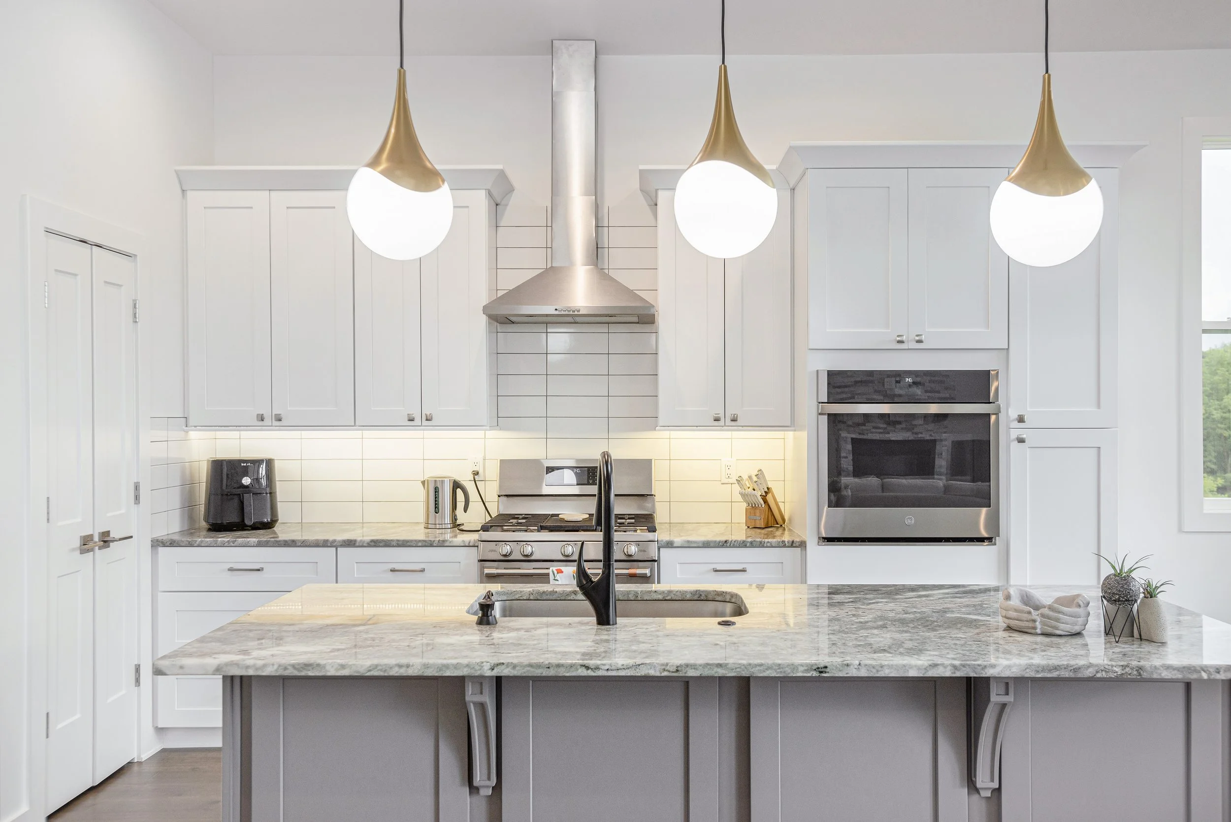 Modern kitchen with white cabinets, stainless steel oven, range hood, and pendant lights over a marble-topped island counter.