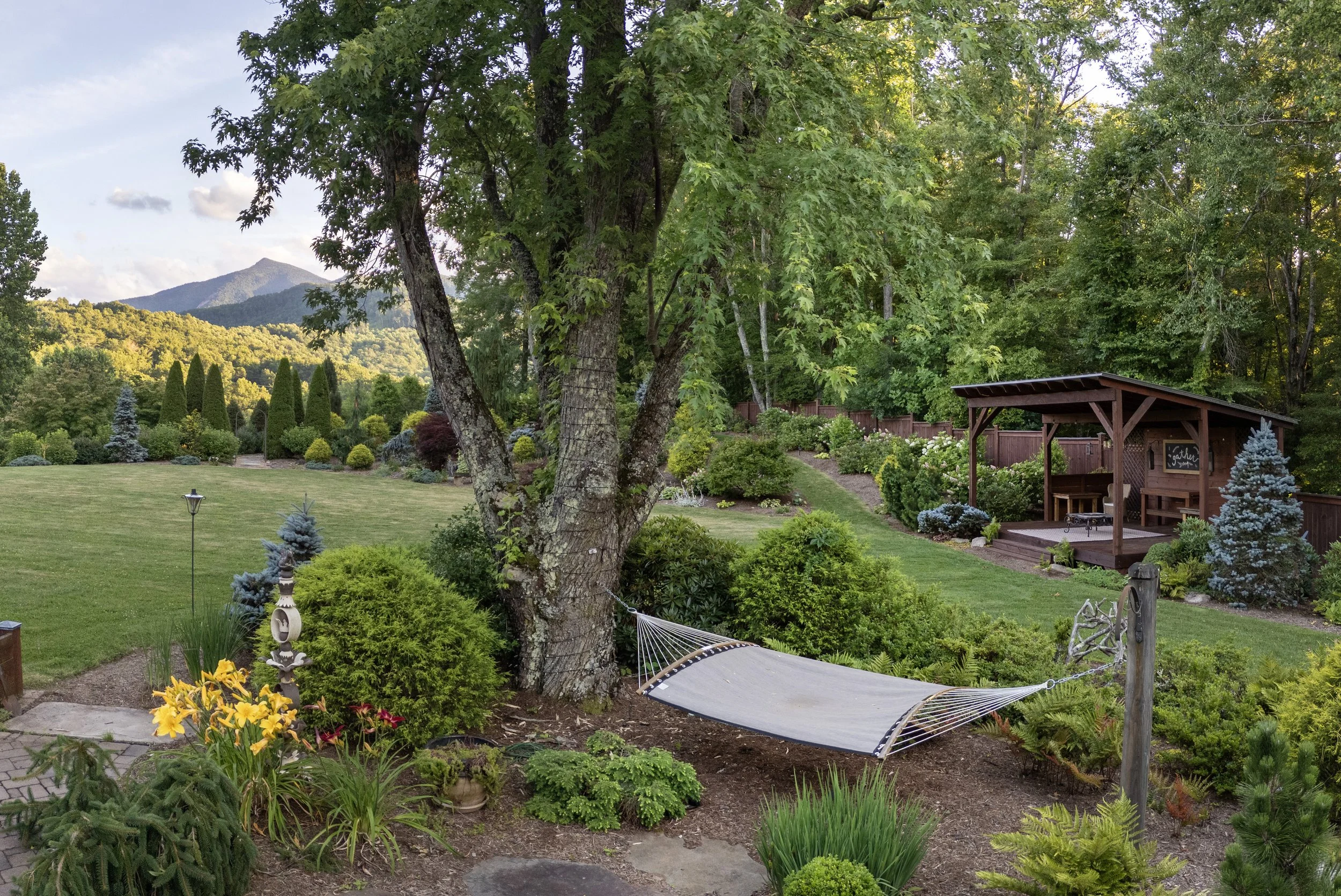 A lush backyard garden with a large tree in the foreground, a green lawn, colorful flowers, shrubs, a hammock, a wooden structure with a roof, and rolling hills and a mountain in the background under a partly cloudy sky.