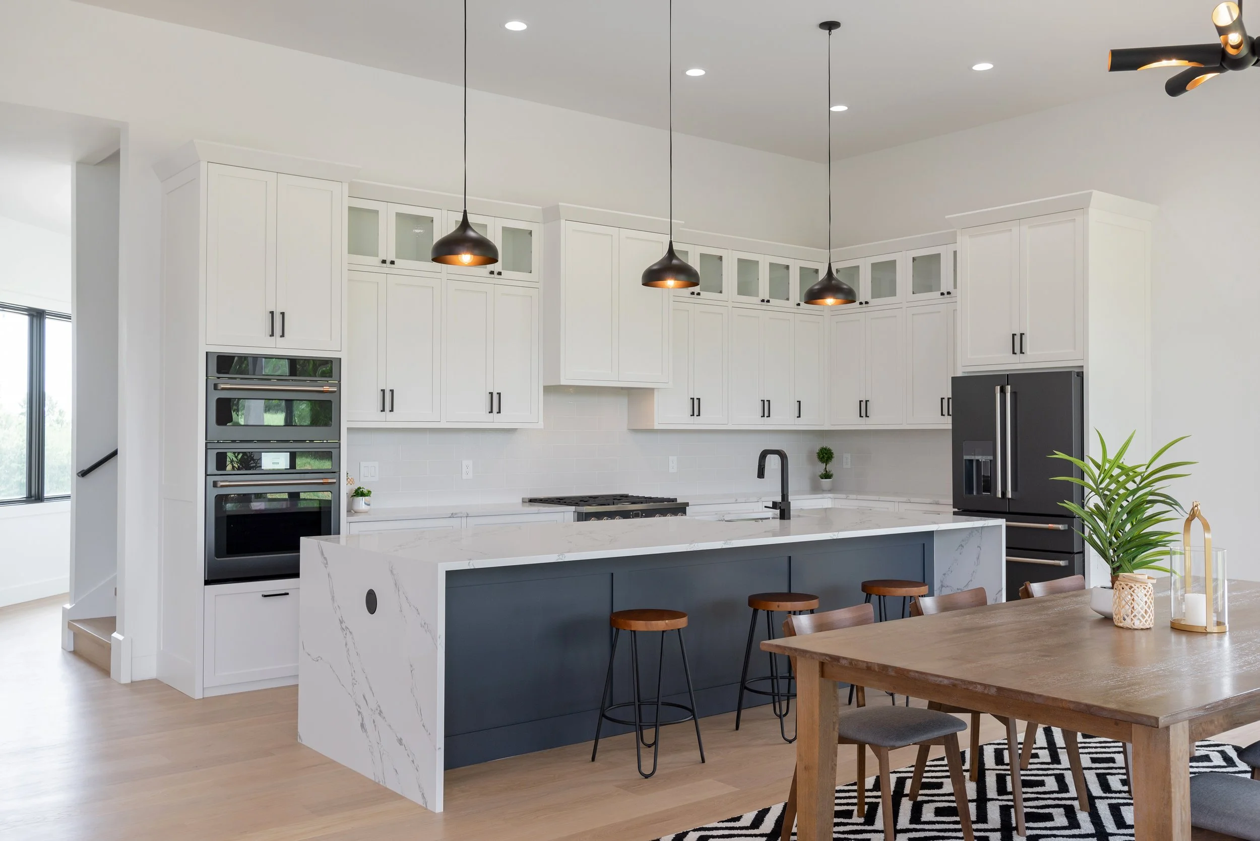 Modern kitchen with white cabinets, black hardware, a marble island, black pendant lights, and a wooden dining table with chairs.