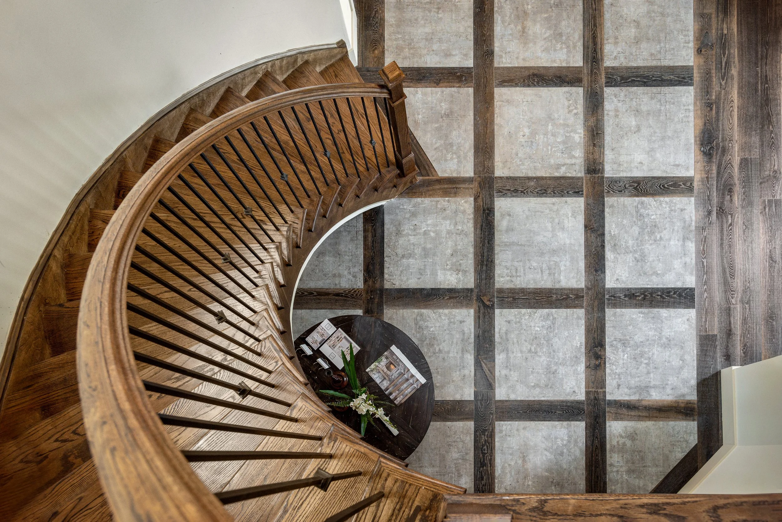 Top-down view of a curved wooden staircase with black railing, overlooking a checkered wooden and stone tile floor, near a small black table with magazines and a plant.