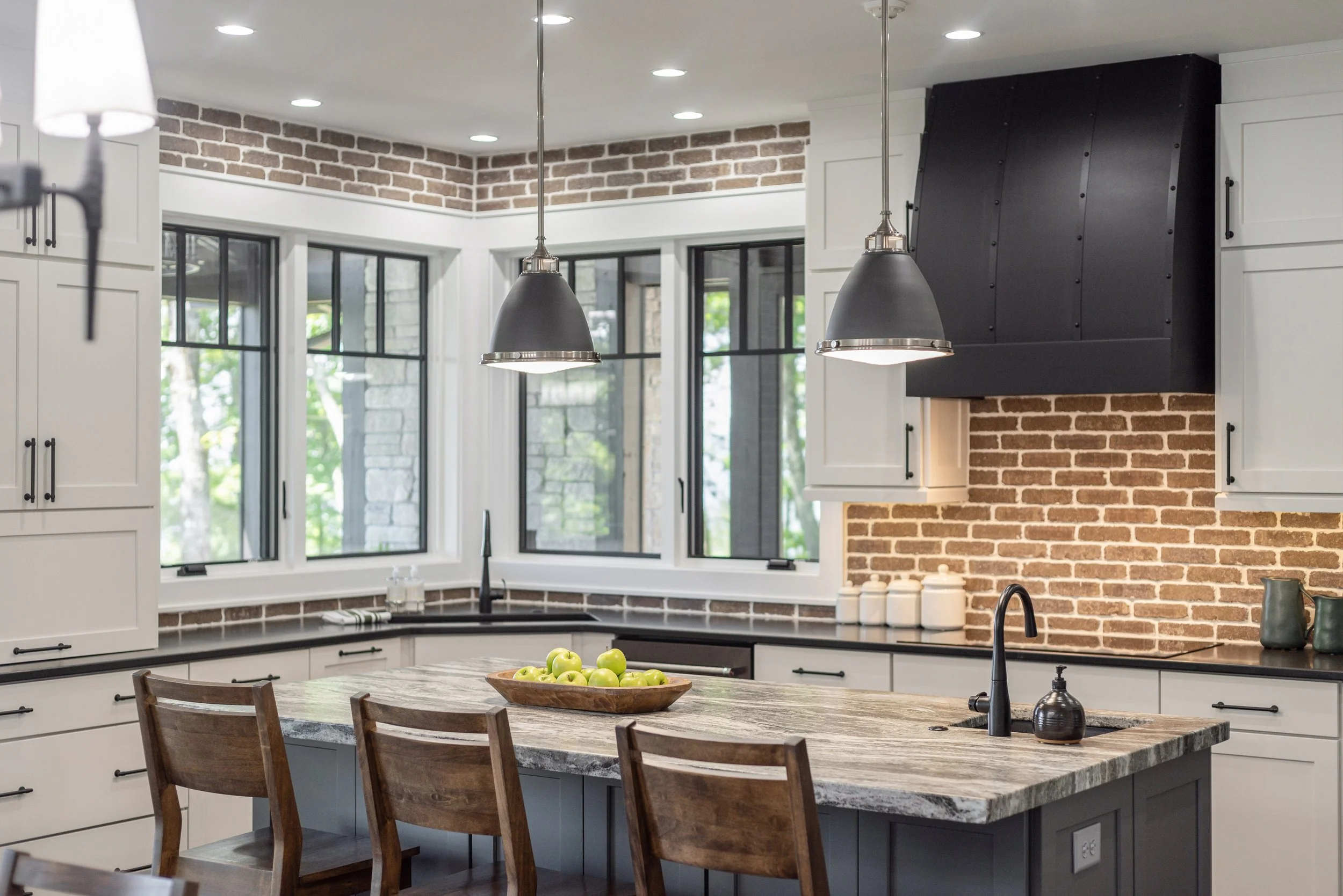 Modern kitchen with white cabinets, black countertops, brick walls, large windows, and hanging black pendant lights over the island with a bowl of green apples.