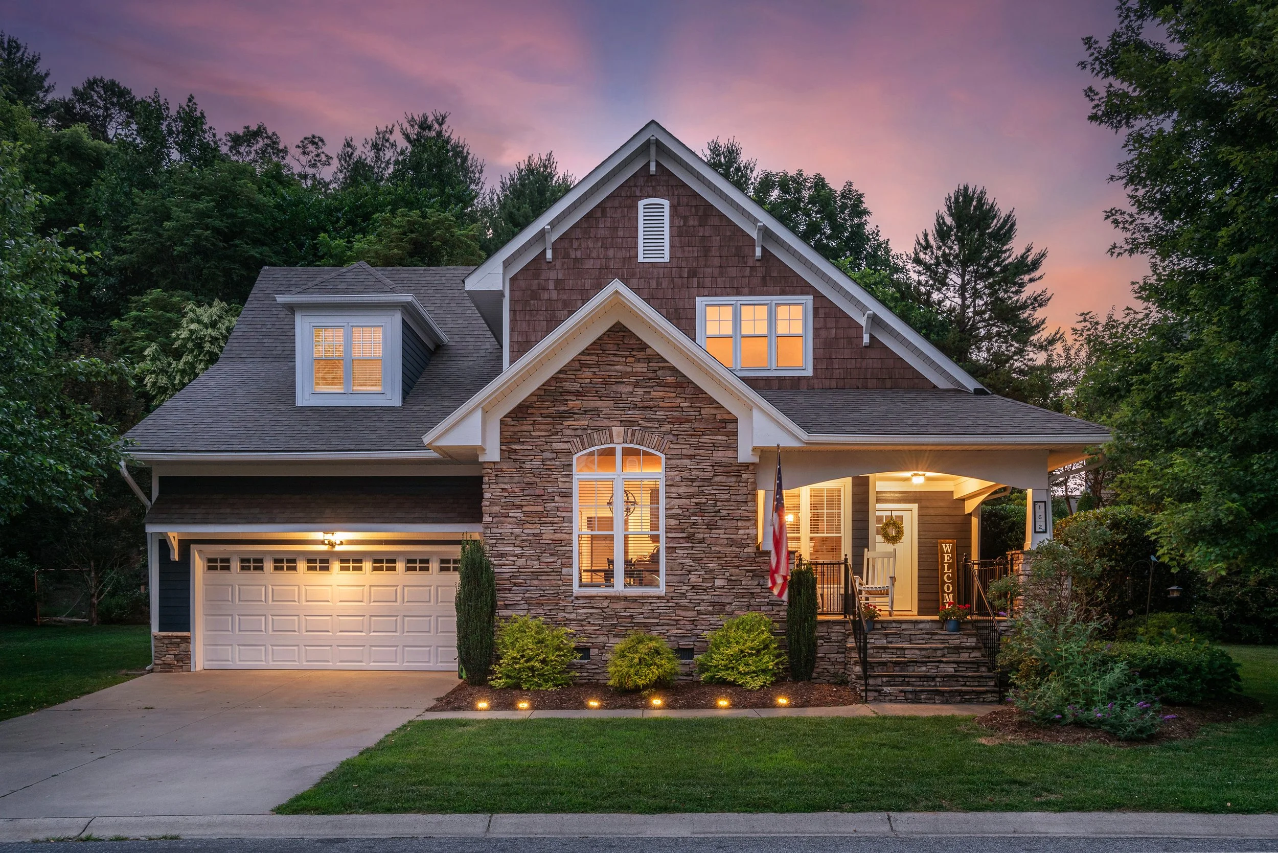 A two-story house with a stone and siding exterior, with illuminated windows, a front porch with a wreath, a welcome sign, and a driveway leading to a closed garage door. The house is surrounded by trees and greenery, and the sky in the background sh