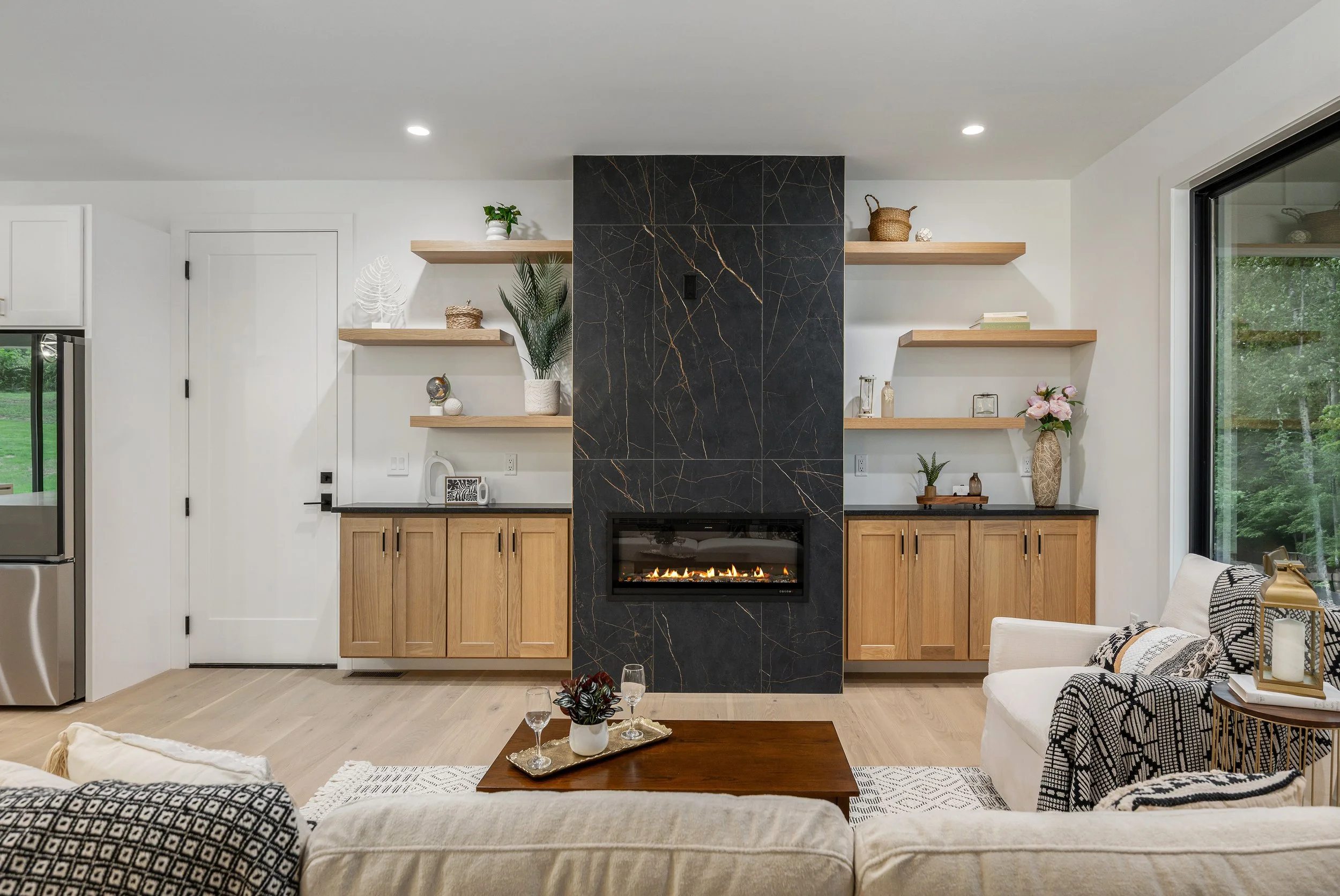 Living room with a modern black marble fireplace, wooden shelves with decor, and large windows showing greenery outside.