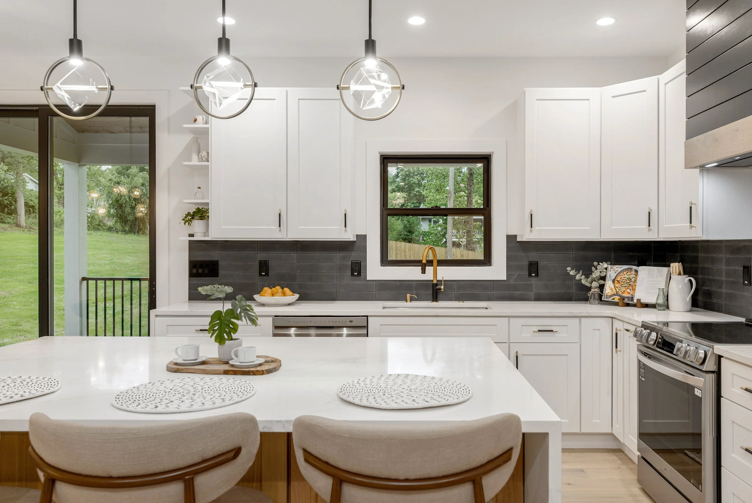 Modern kitchen with white cabinets, black tile backsplash, and a large window overlooking greenery. Three pendant lights hang above a white island with a fruit bowl and a small potted plant. A sliding glass door to the left shows an outdoor yard.