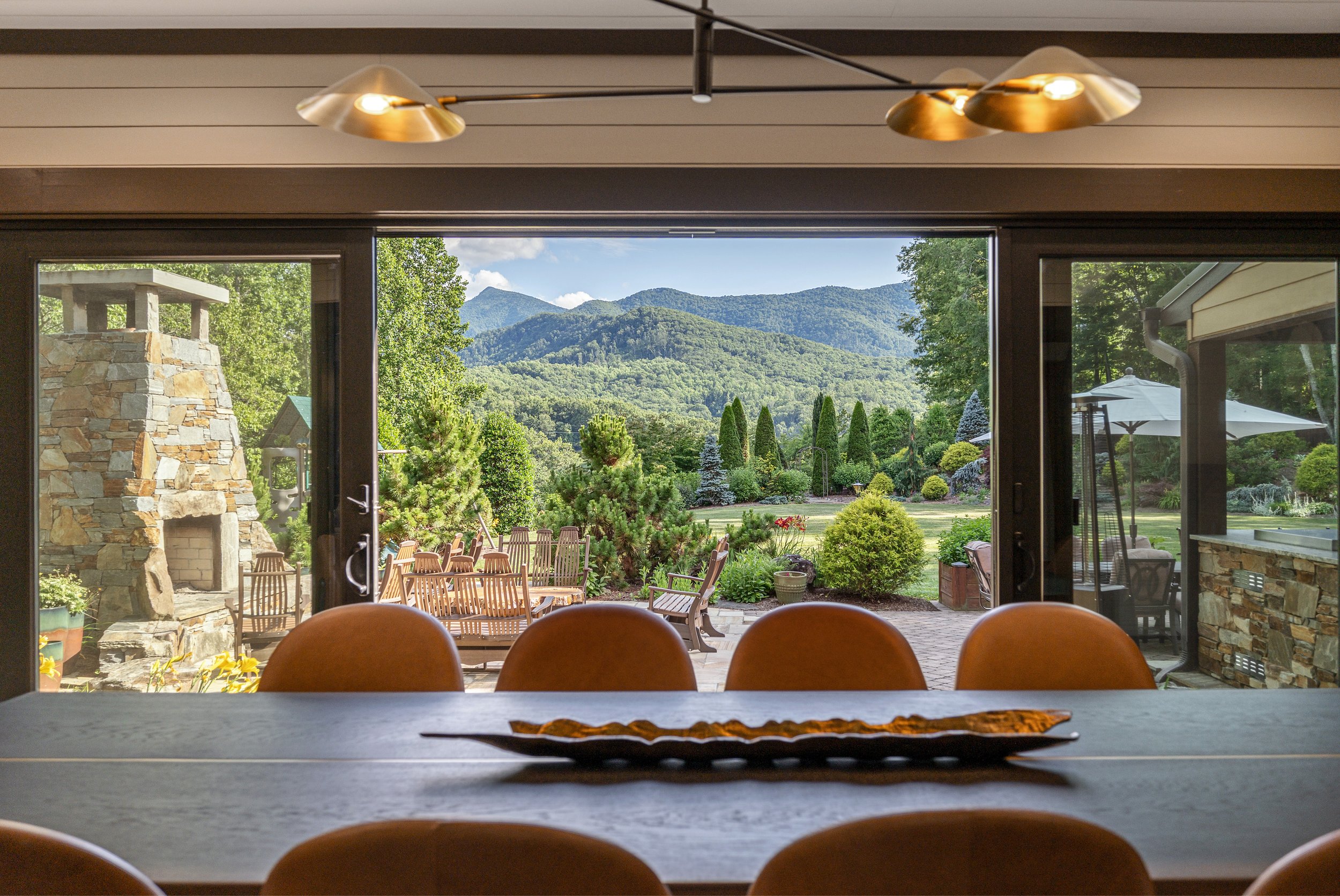 View of a garden and mountains through sliding glass doors from inside a room with a stone fireplace and a wooden dining table with chairs.