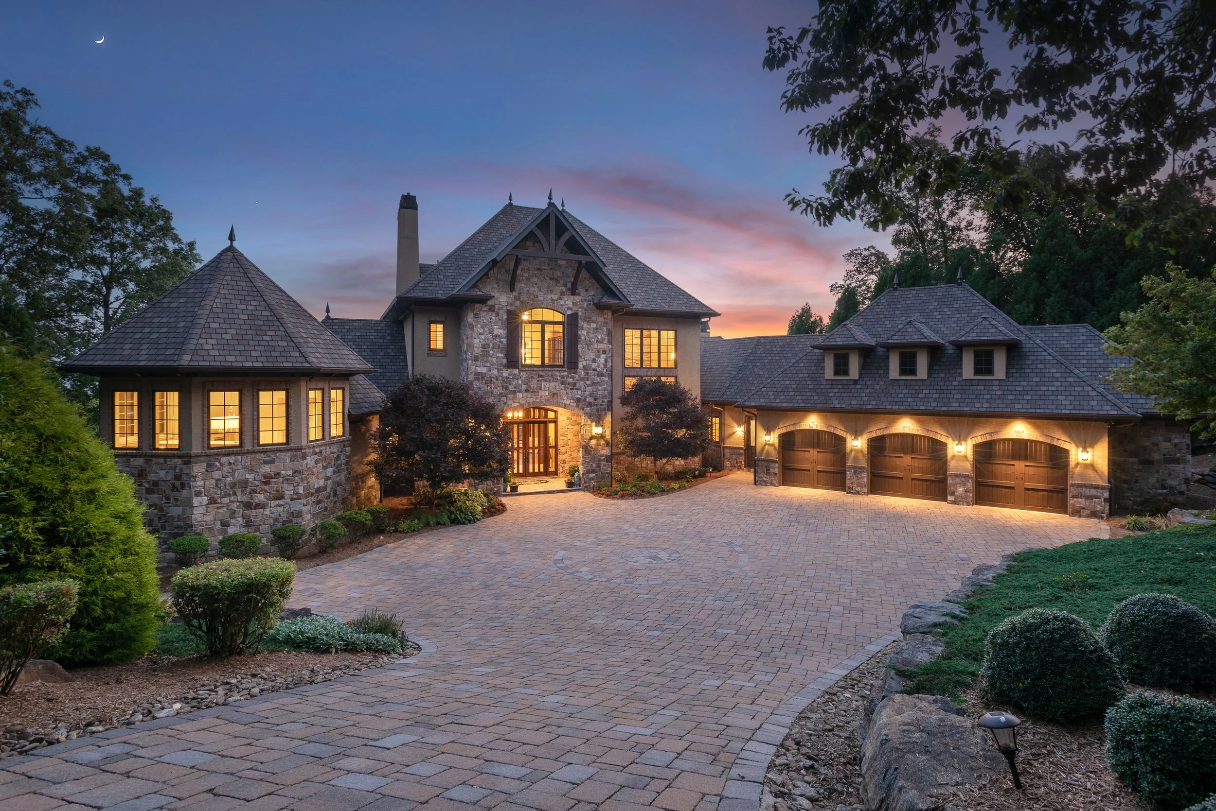 A large house with stone walls and multiple windows, illuminated from within, during twilight. The house features several peaked roofs and a driveway paved with bricks, surrounded by lush greenery and trees.