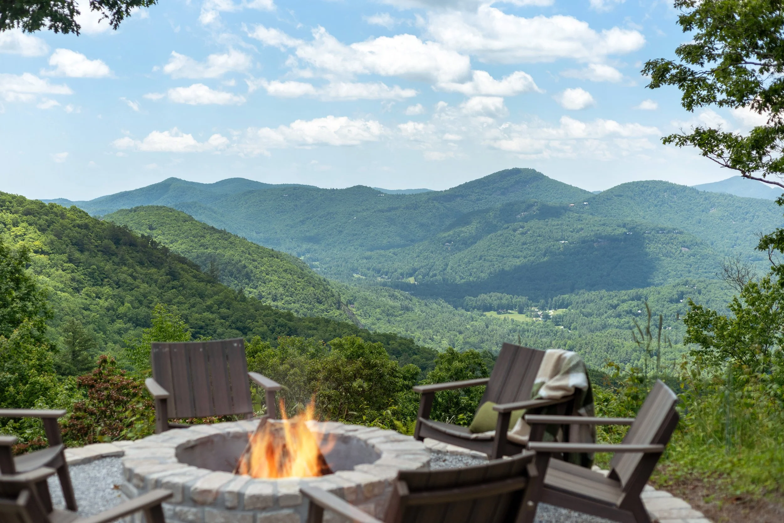 A scenic view of green mountains under a cloudy sky, framed by trees, with a patio area in the foreground featuring outdoor chairs surrounding a fire pit with a small fire.