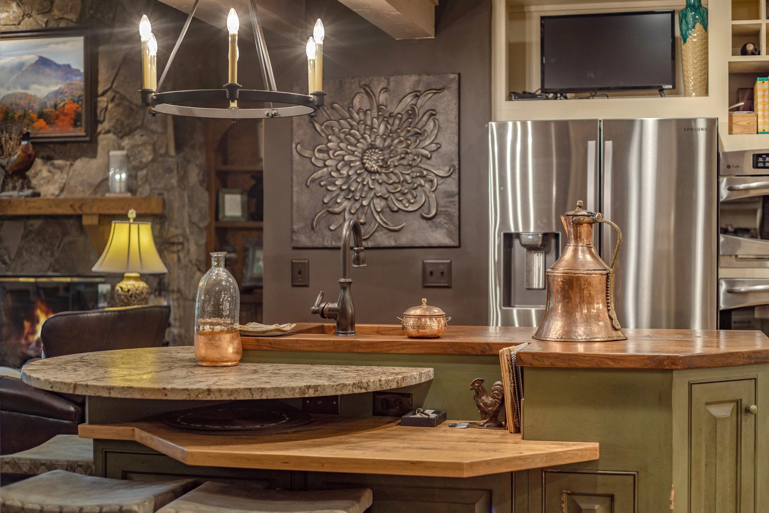A cozy kitchen and dining area with a stone wall, decorative art, a chandelier, a stainless steel refrigerator, a television, and various decorative items including copper pitchers and a table lamp.