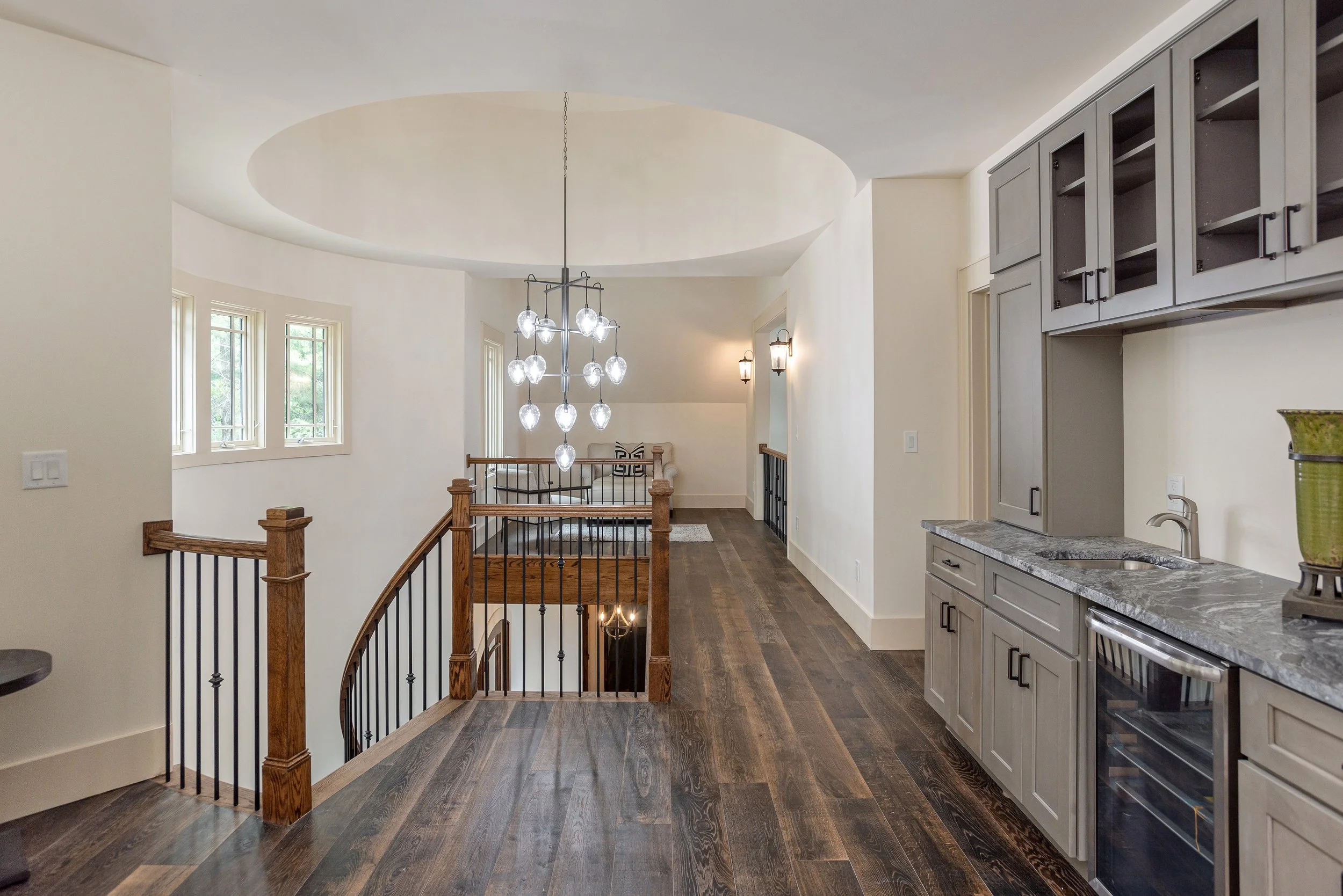 Interior view of a modern staircase landing with hardwood floors, a contemporary chandelier, large windows, and gray kitchen cabinets with a granite countertop.