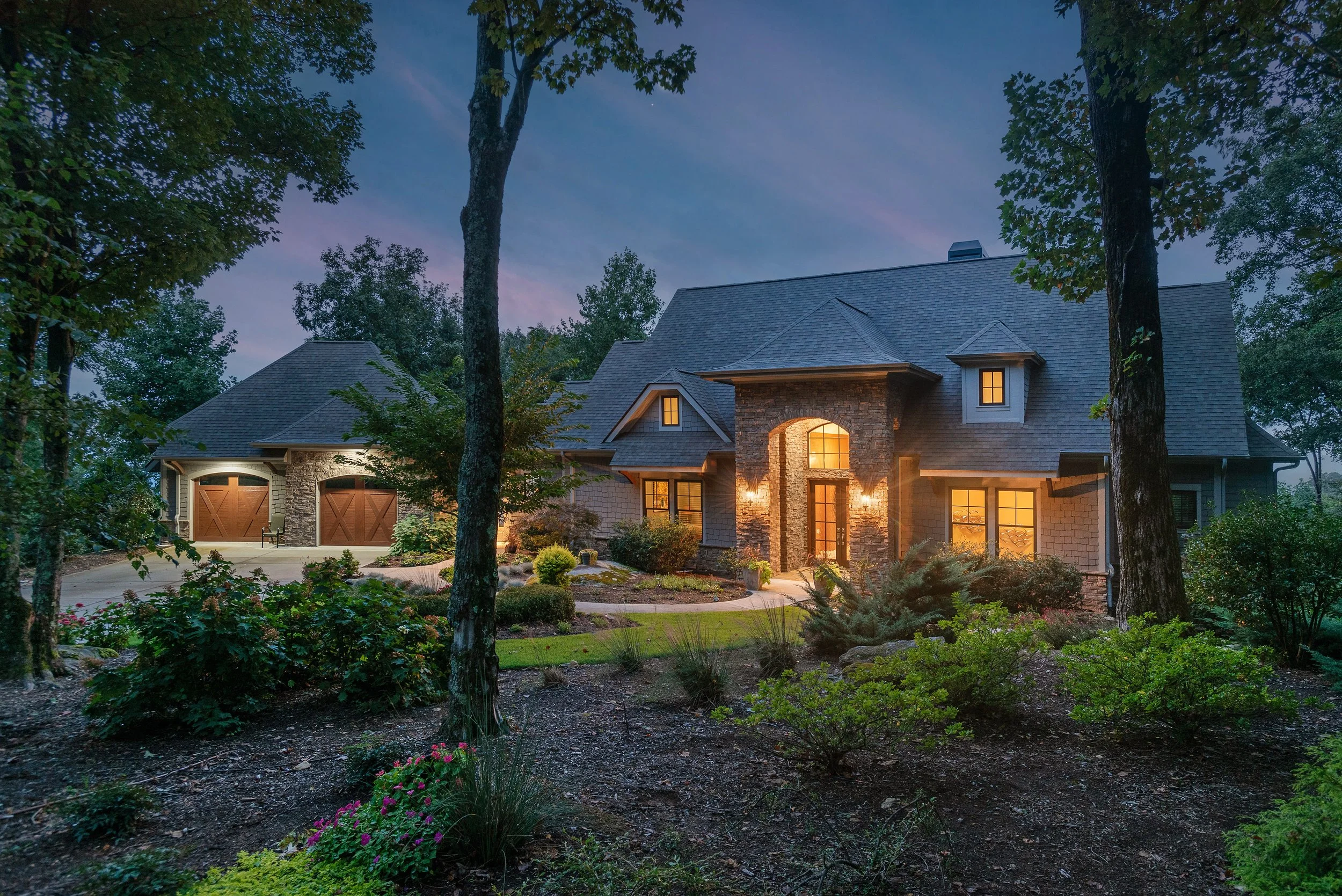 A large house with stone and siding exterior, multiple peaks on the roof, illuminated windows, surrounded by trees and landscaped garden, with evening sky in the background.