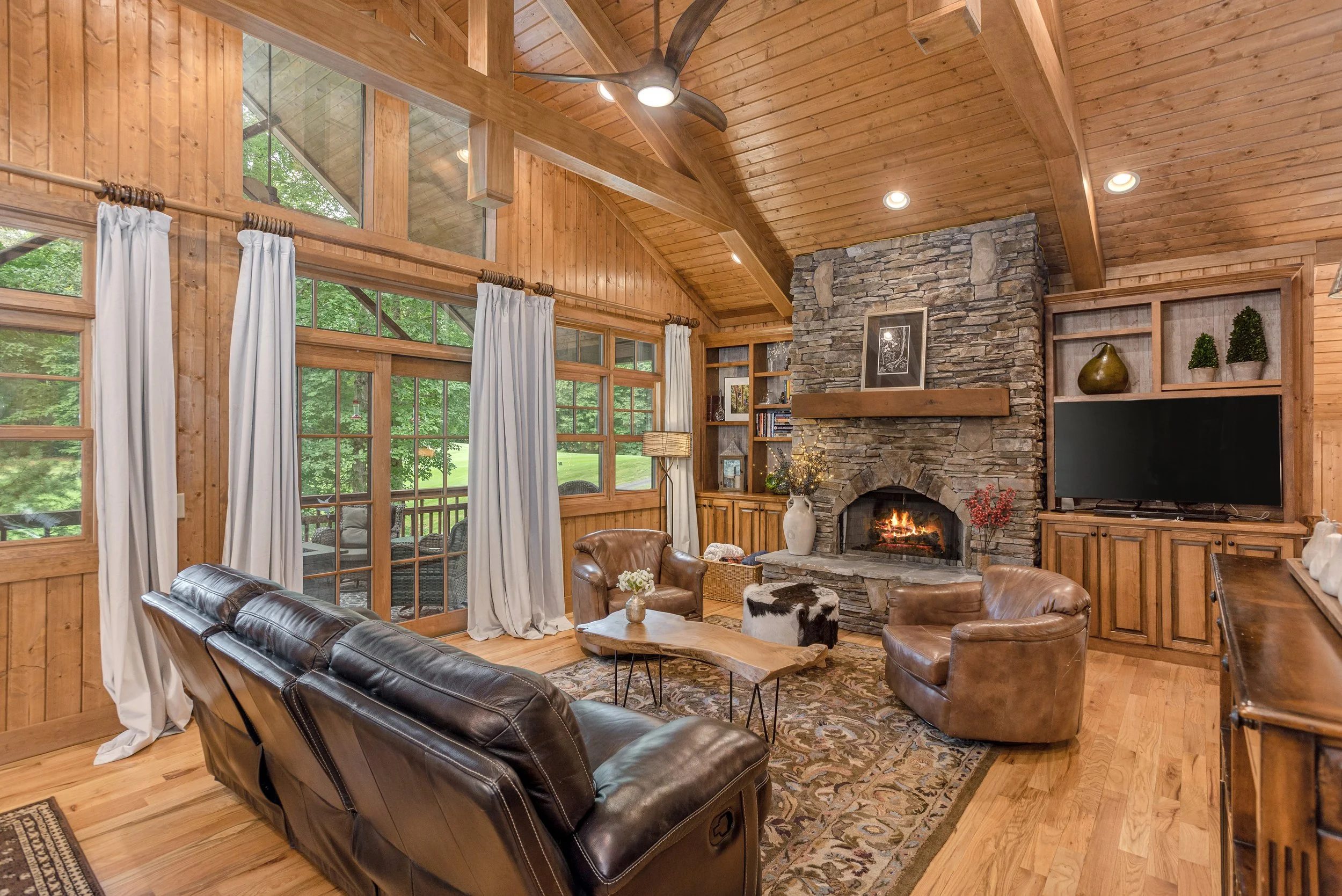 Living room with wood-paneled walls, large windows with white curtains, a stone fireplace with a framed picture on the mantle, leather chairs, a rug, and a ceiling fan.