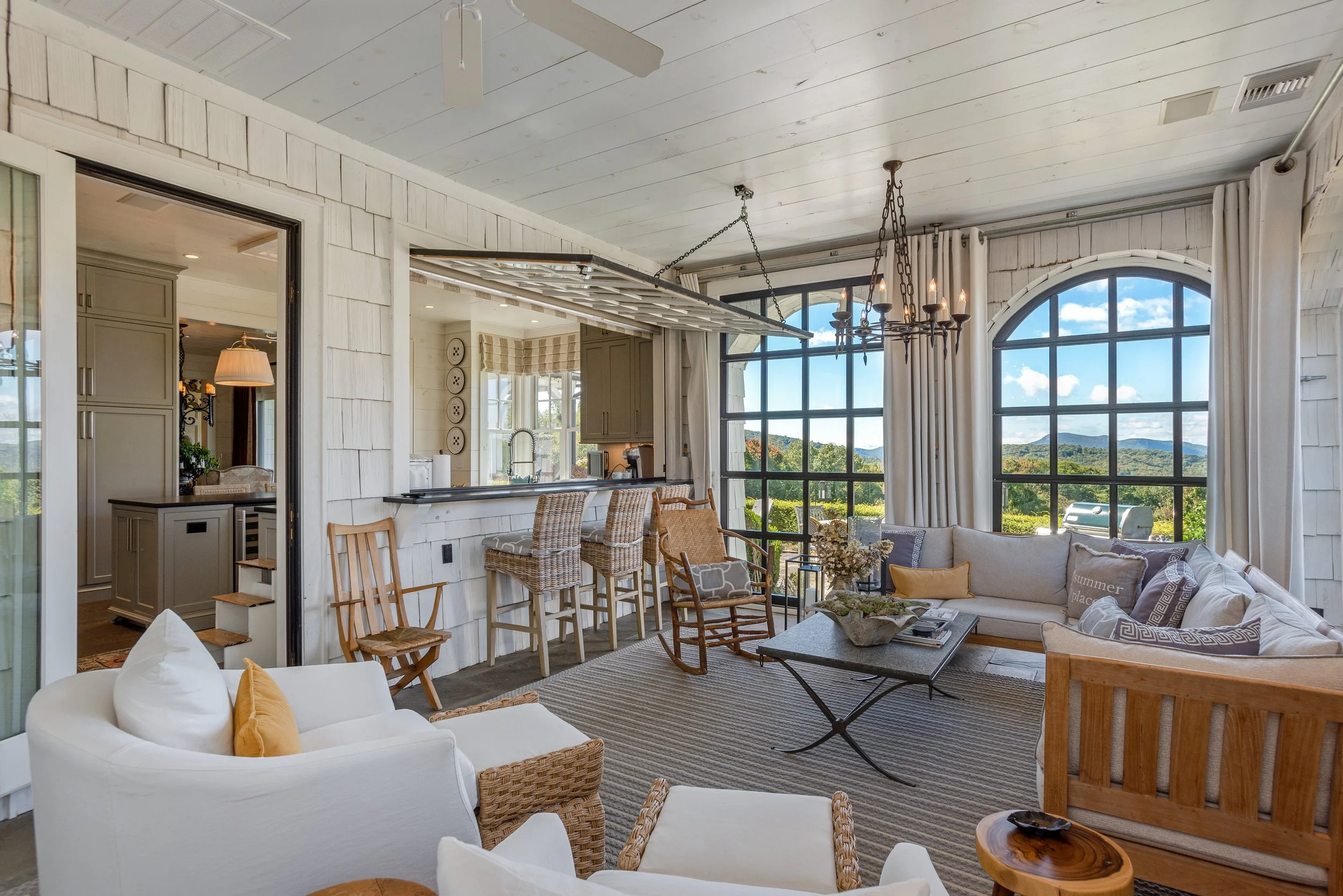 A bright living room with large bay windows showcasing a scenic mountain view, decorated in a coastal style with white shiplap walls, wicker chairs, a white upholstered sofa with yellow pillows, and a coffee table with decorations. The ceiling has a 
