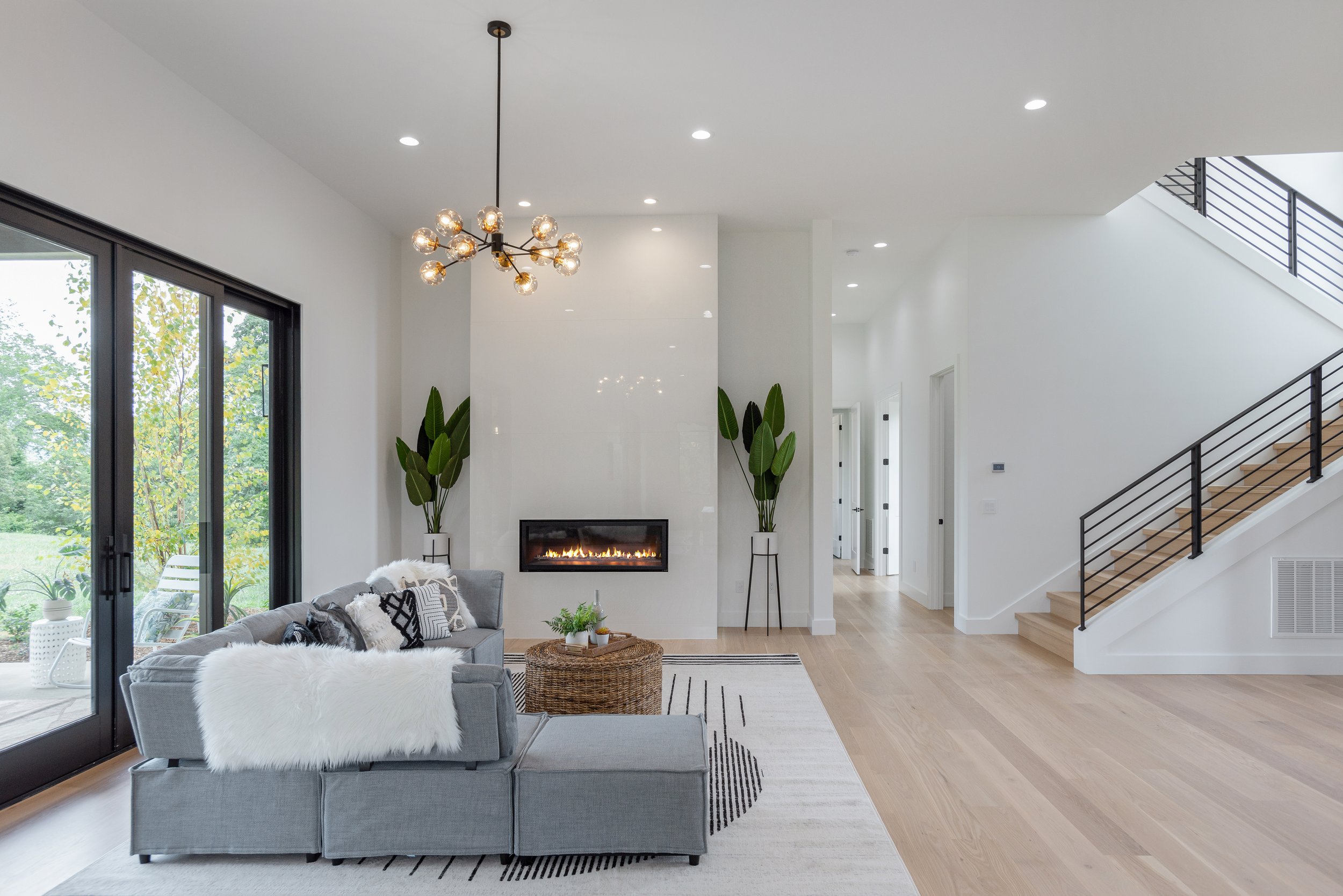 Modern living room with gray sofa, fur throw blanket, patterned pillows, and round wicker coffee table in front of a fireplace, large planted leaves on either side of fireplace, black sliding glass door to patio, staircase with black railing, and con