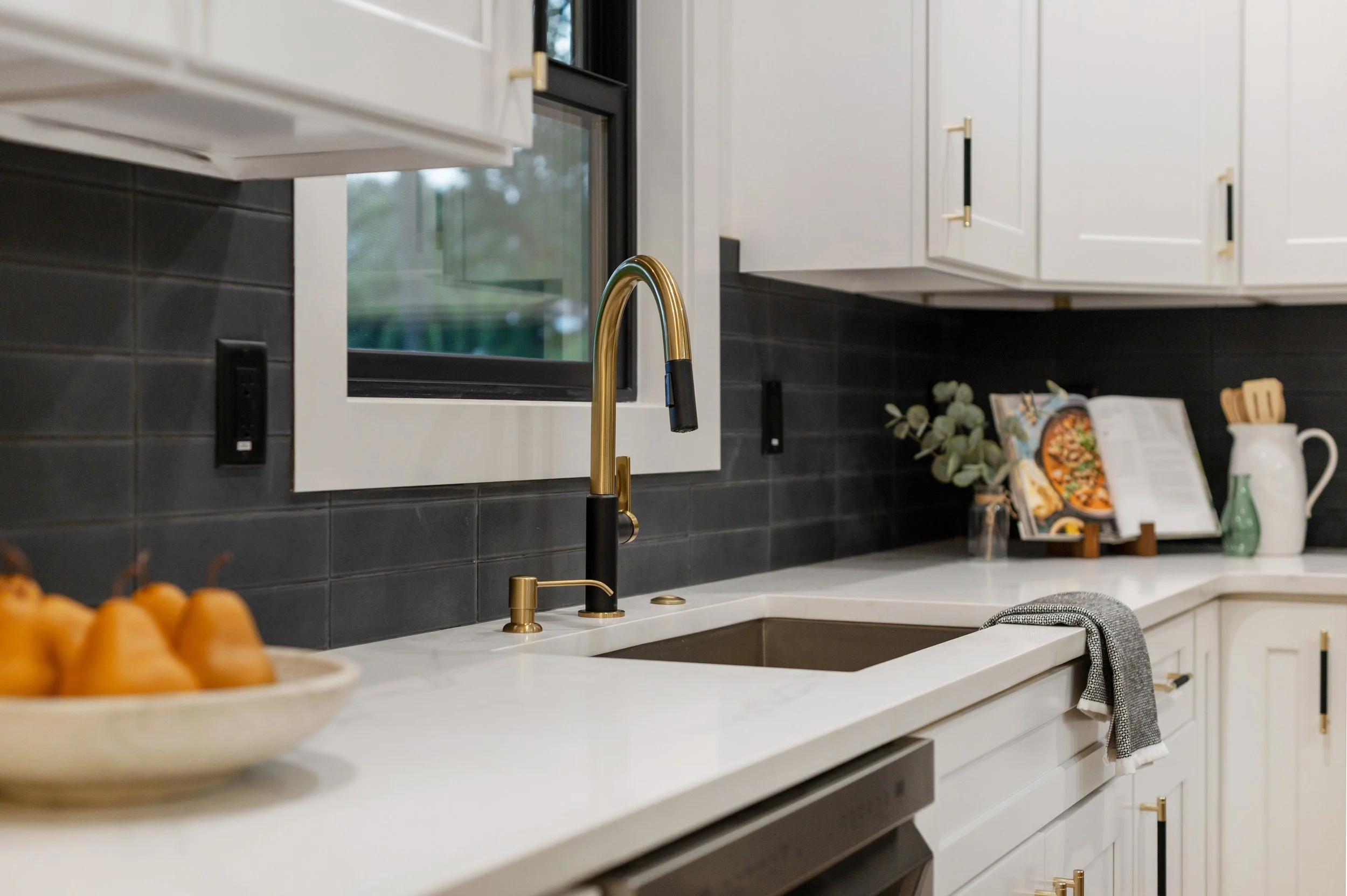 Kitchen with white cabinets, black backsplash tiles, a gold kitchen faucet, and a window above the sink. A bowl of oranges is partially visible in the foreground.