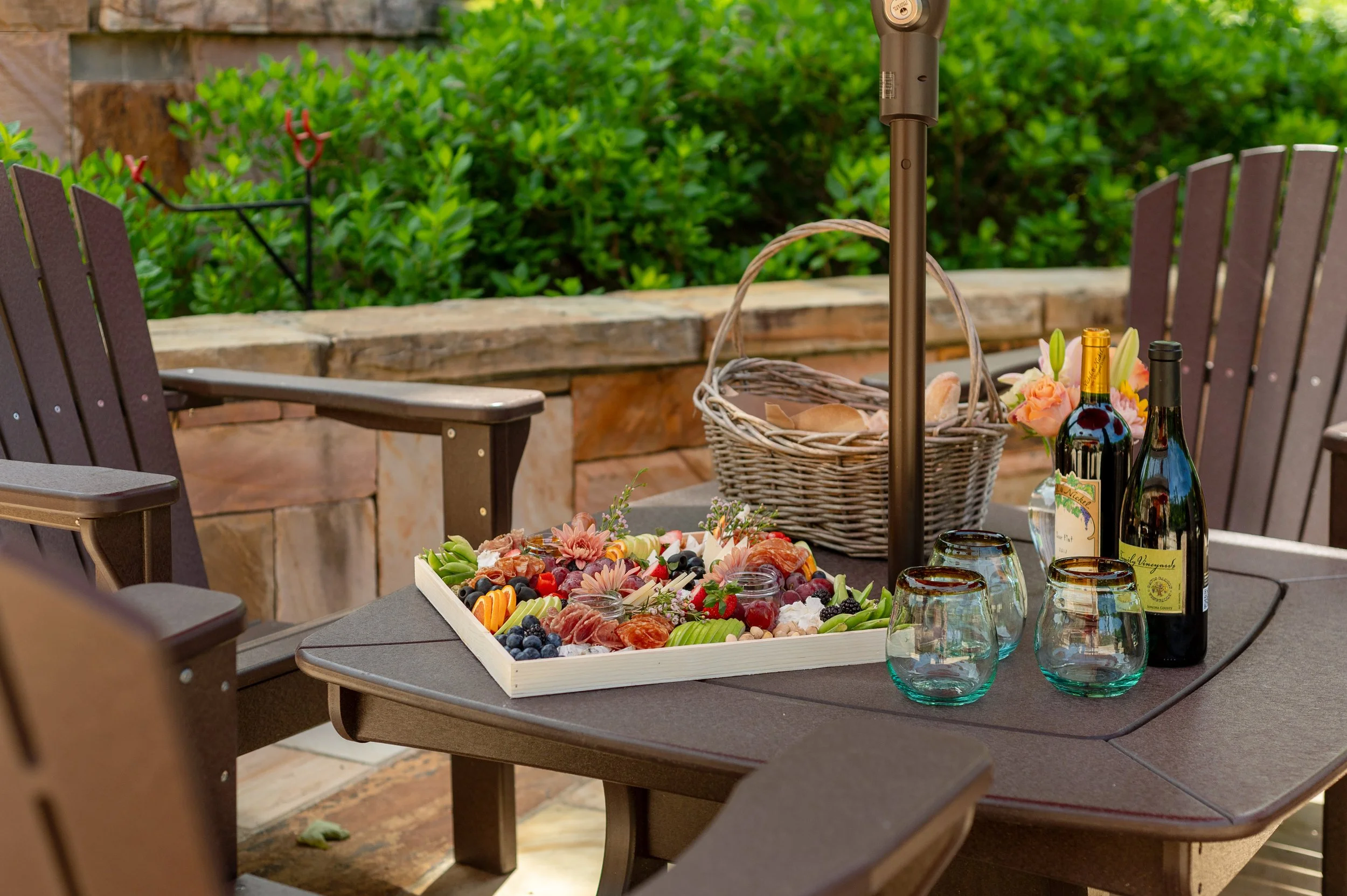 An outdoor patio with a table set for a gathering, featuring a tray of fresh fruits, two bottles of wine, and empty glasses, surrounded by wooden chairs and greenery in the background.